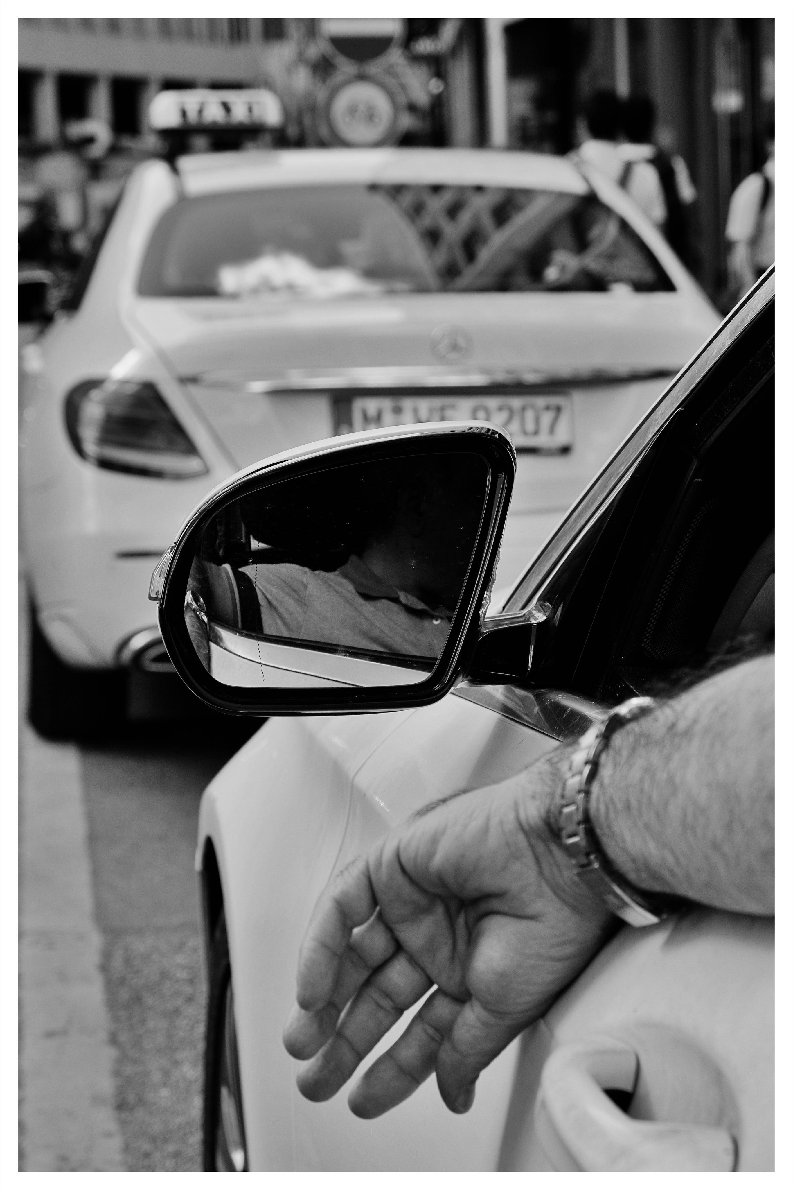 Black-and-white street scene with a close-up of a car’s side mirror and an arm resting out the window in the foreground; a taxi is stopped ahead with pedestrians and street signs blurred in the background.