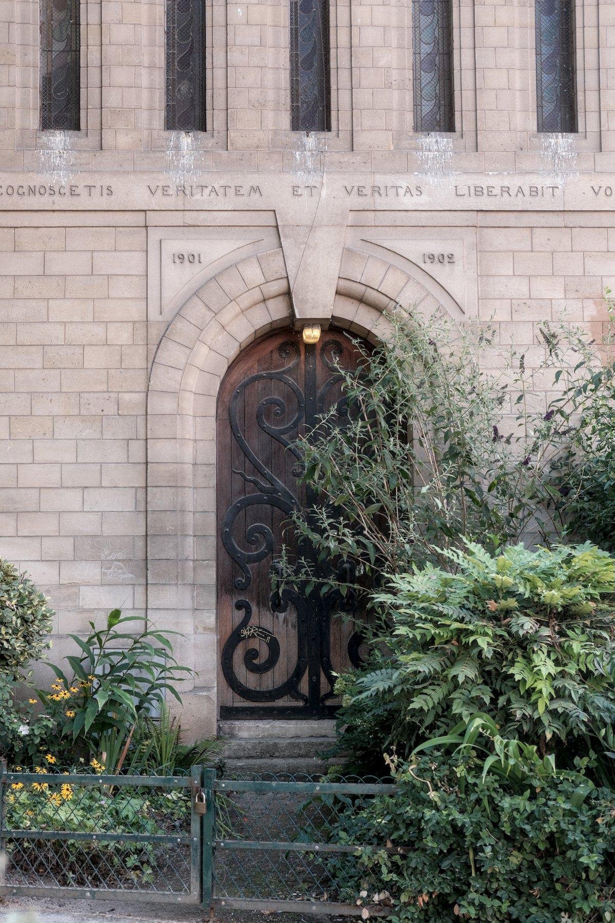 Photo verticale. Derrière une petite barrière verte de jardin parisien, une grande porte en bois au sommet en arche, décorée de fer forgé plaqué dessus, dans un mur en pierre régulières taillées, beige