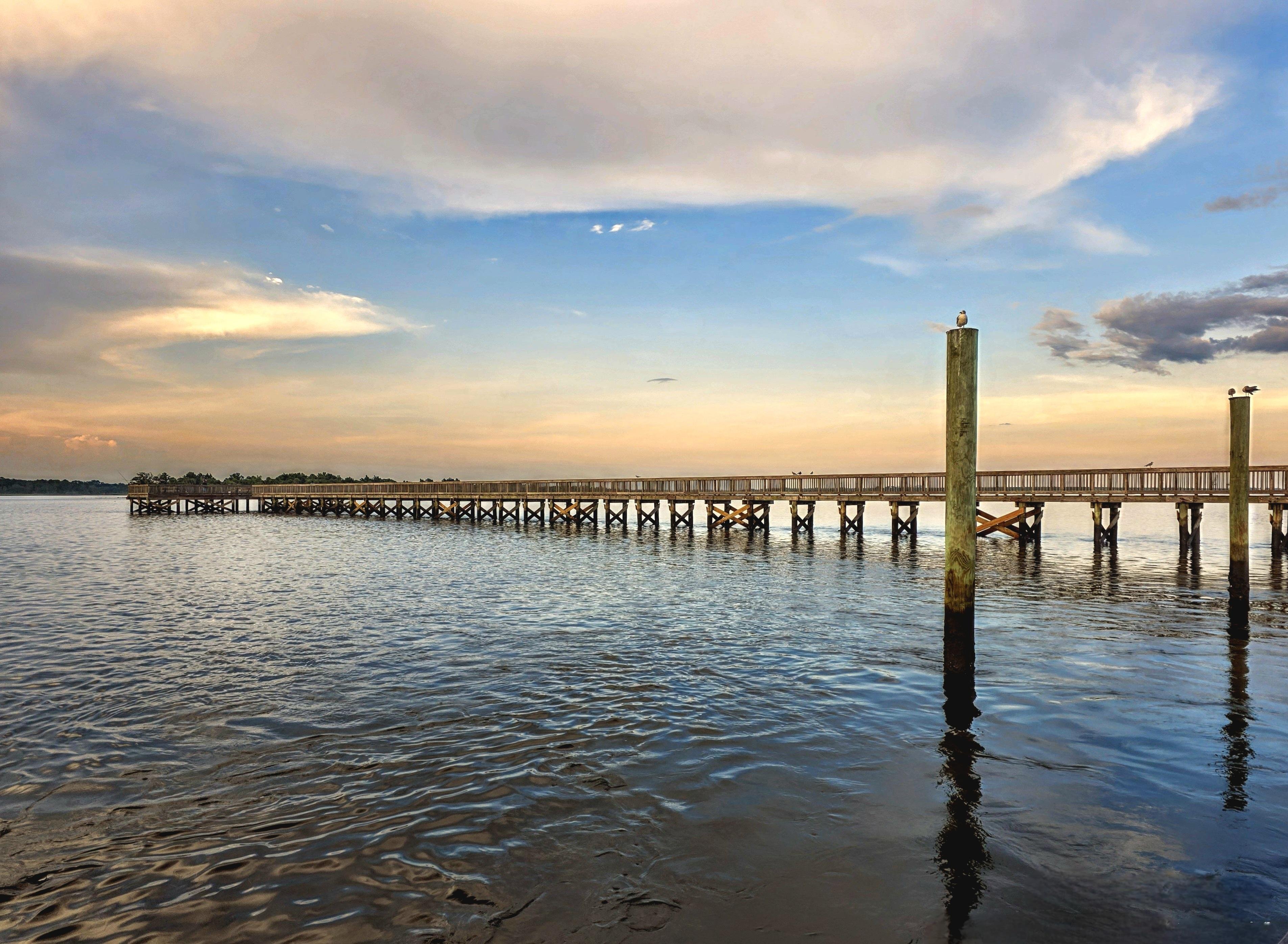 Wooden pier extending across a calm body of water during sunset, with soft orange and blue hues in the sky. A few birds perch on the pier and nearby posts, and the surface of the water reflects the colors of the sky.