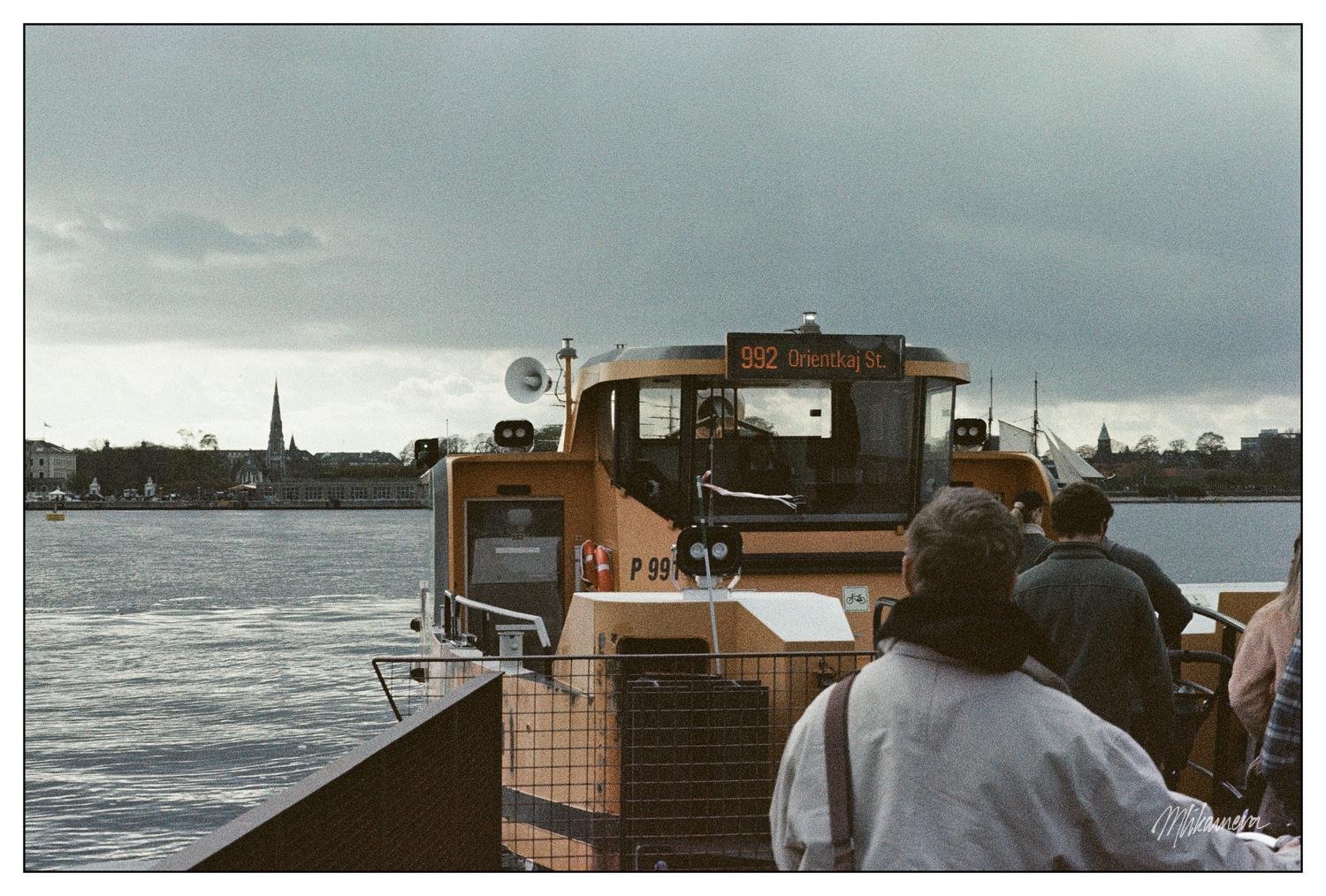 A ferry with the destination "992 Orientkaj St." displayed on its front, docked at a pier. Several people are waiting to board, with their backs to the camera. The background features a body of water and a silhouette of a town, including a church spire under a cloudy sky.