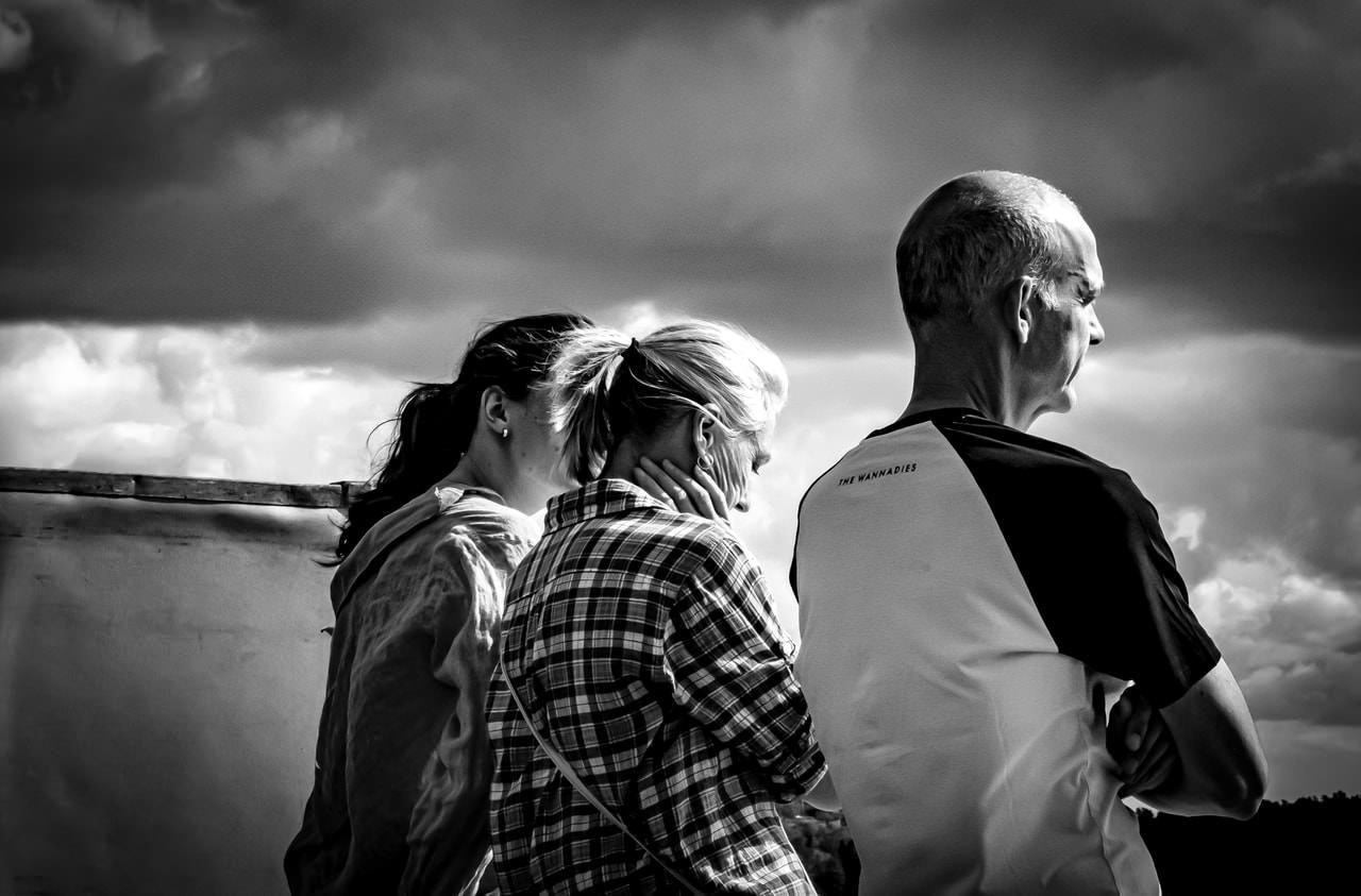 Three adults stand closely together, viewed from the side, looking towards the sky. The person on the left has long dark hair tied back, the middle person has light hair in a ponytail and is wearing a plaid shirt, and the person on the right has short hair and is wearing a two-toned shirt. The background features a cloudy sky with dramatic contrasts in light and shadow. The image is in black and white.