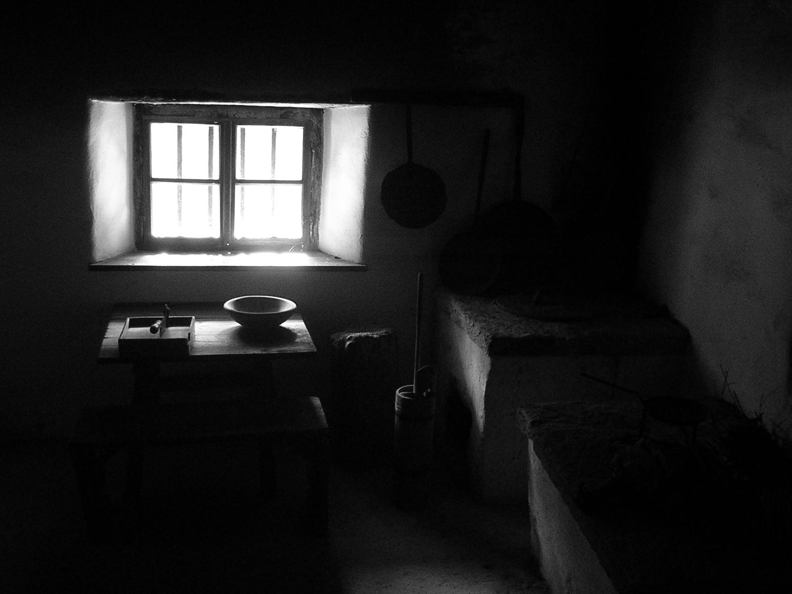 The image shows a dimly lit room with a small window letting in light. In the room, there is a table with a bowl and some objects on it. Kitchen utensils such as pans are hanging on the wall, and there appears to be a hearth or stove area to the right. The scene gives an old-fashioned or rustic kitchen vibe.