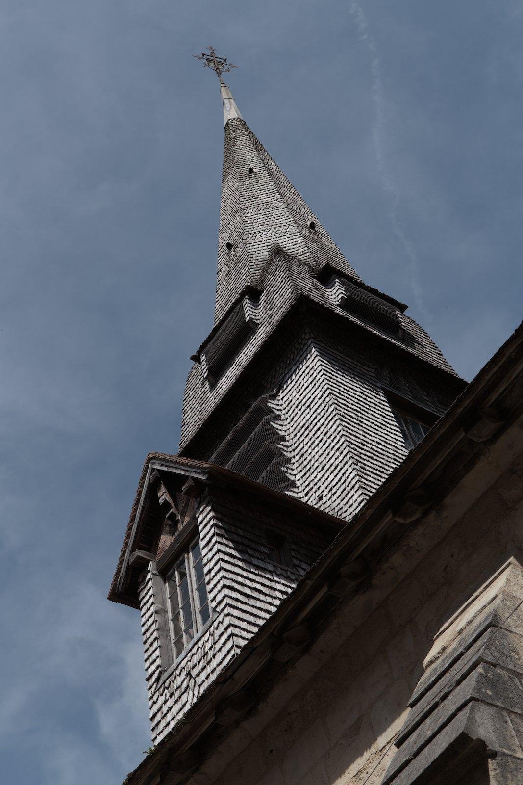 Photo format portrait en contre plongée montrant le clocher d’une église, couverte de petite tuile (peut être en ardoise) donnant un effet très texturé, grâce aux ombres dû au soleil, sur fond de ciel bleu avec des nuages laiteux