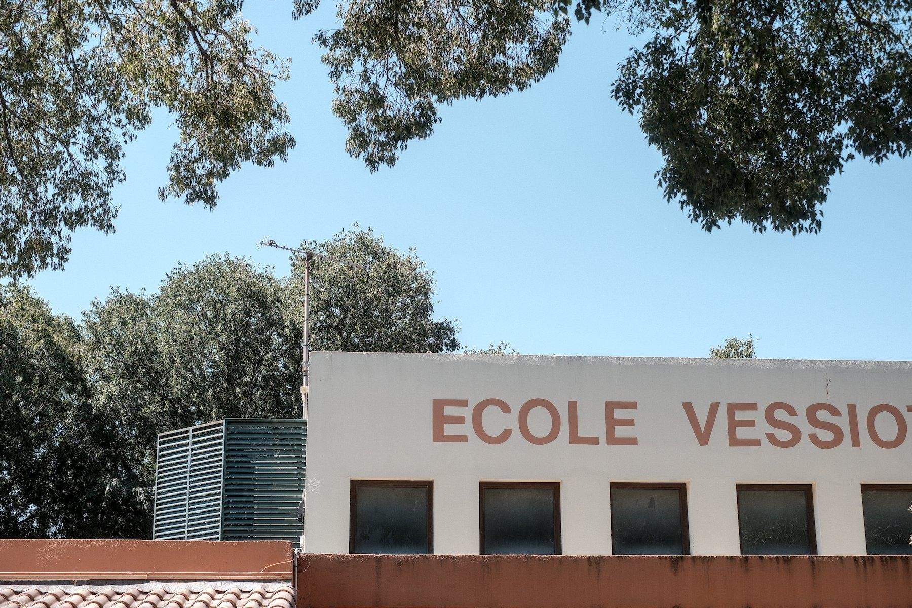 Photo horizontale. Vue sur le haut d’une école, bien rectangulaire, beige, les lettre ECOLE VESSIO peintes en ocre dans une typo simple au possible. À gauge, une espèce de cage de service verte, qui semble protéger soit l’unité extérieur d’une clim, soit une armoire électrique. En haut de l’image, des feuillages d’arbre cadre le tout