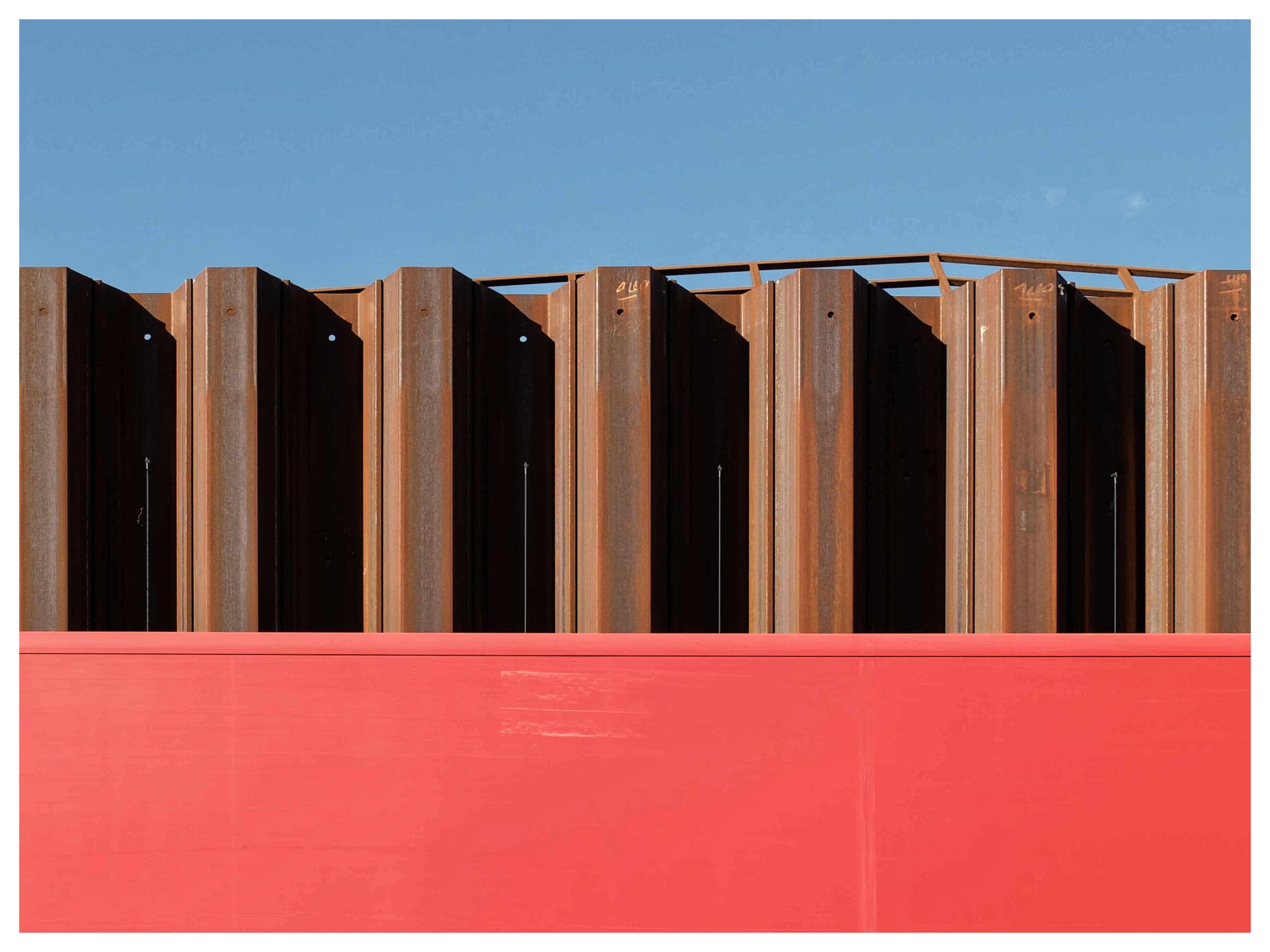 Rows of vertical, rusted metal beams protrude above a bright red horizontal surface, against a clear blue sky.