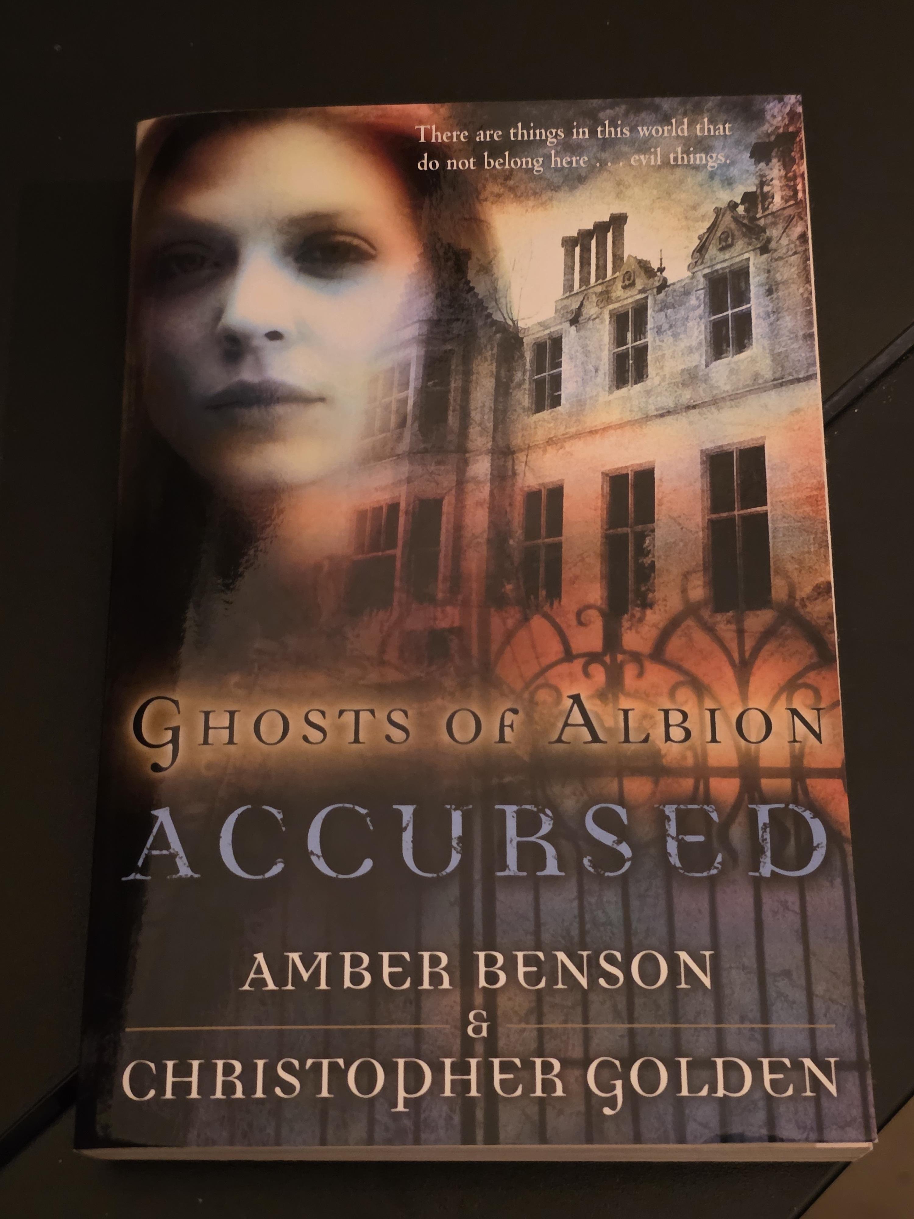 The cover of the supernatural novel "Ghosts of Albion: Accursed" by Amber Benson and Christopher Golden presents a haunting, Victorian-inspired scene. A ghostly, pale face of a woman fades into the upper left corner, her expression somber and ethereal. She overlooks a looming, dilapidated stone manor house that glows with an eerie orange light from within, suggesting it is either on fire or magically charged.
In the foreground, a dark, ornate wrought-iron gate stands closed, separating the viewer from the mist-shrouded estate. The title "GHOSTS OF ALBION" is written in an elegant, serif gold font, while the word "ACCURSED" appears below in a large, distressed blue-grey typeface. This book is the first in a series originally based on a BBC animated web drama, set in a mystical 1838 London where a brother and sister must protect England from ancient, malevolent forces.
