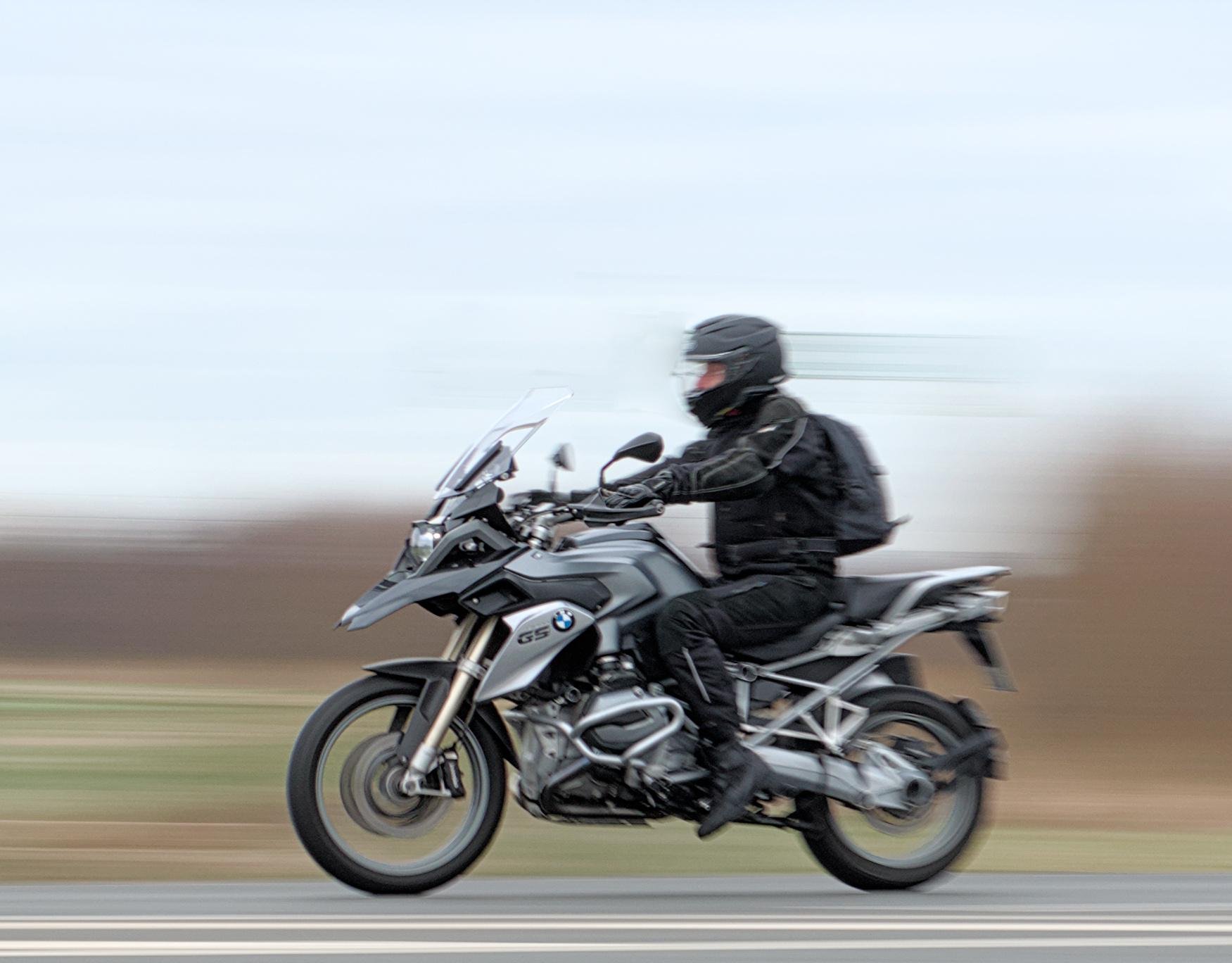 The image shows a person riding a motorcycle at speed on a road. The motorcycle has a sleek, modern design, and the rider is wearing full protective gear, including a helmet. The background is blurred, indicating motion.