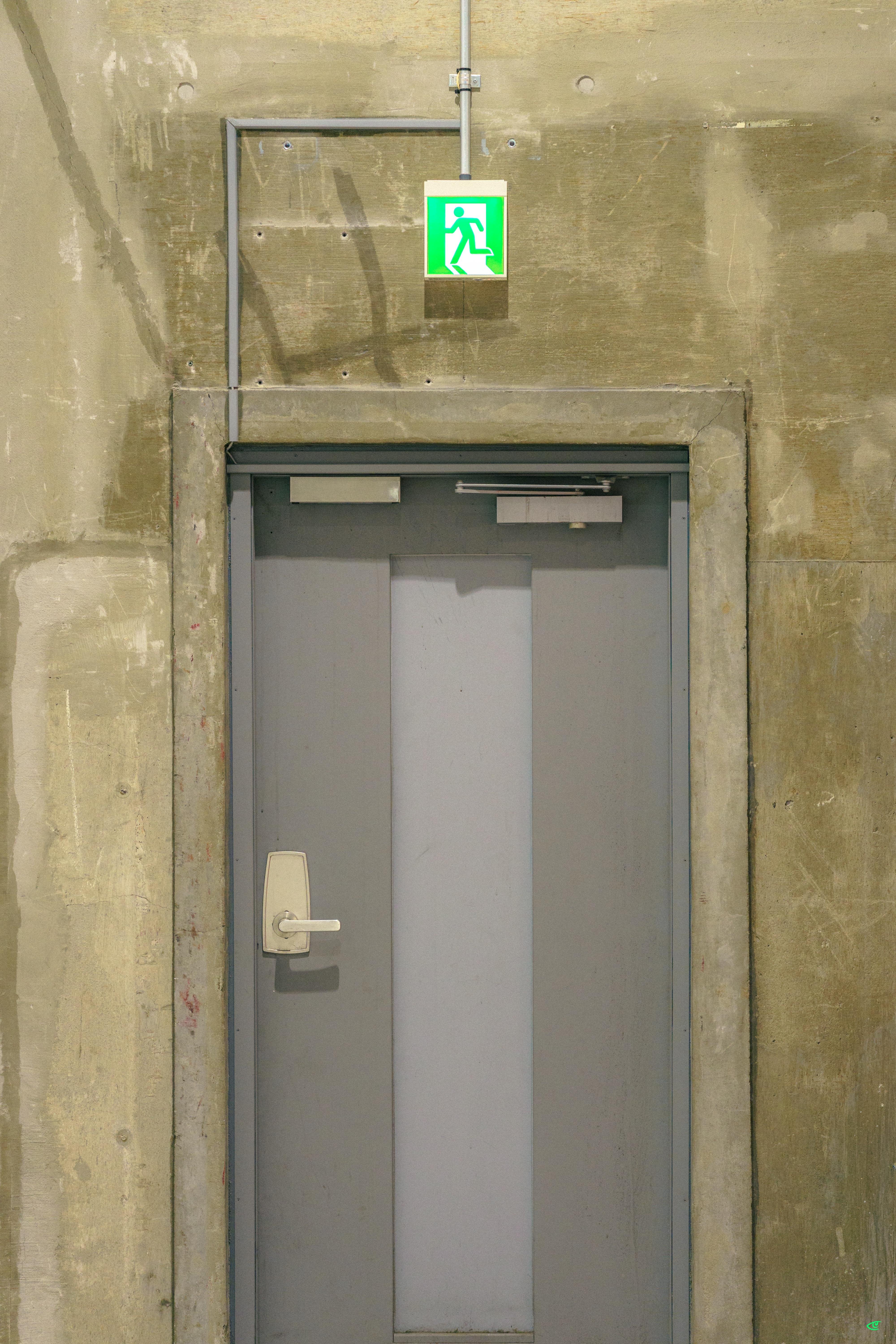 Gray metal emergency exit door set in a concrete wall, with a narrow vertical window panel, a lever handle on the left, and a green illuminated running-man exit sign mounted above.