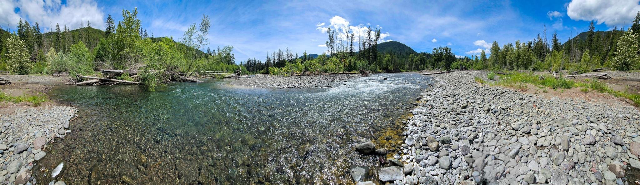 Panoramic view of a river flowing over smooth stones, surrounded by dense green trees and shrubs. A clear, blue sky with scattered clouds arches over the scene. Hills with tree-lined slopes rise in the background. The riverbank is lined with stones and logs, creating a natural, serene landscape.