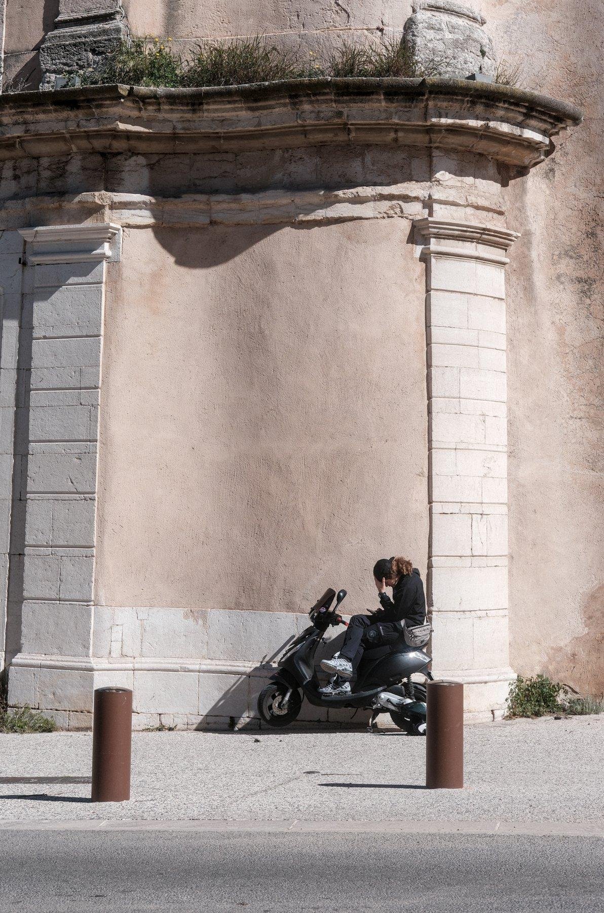 Photo verticale. Une personne avec sa casquette portée à l’envers, consulte son téléphone assis sur son scooter noir, en se tenant le front