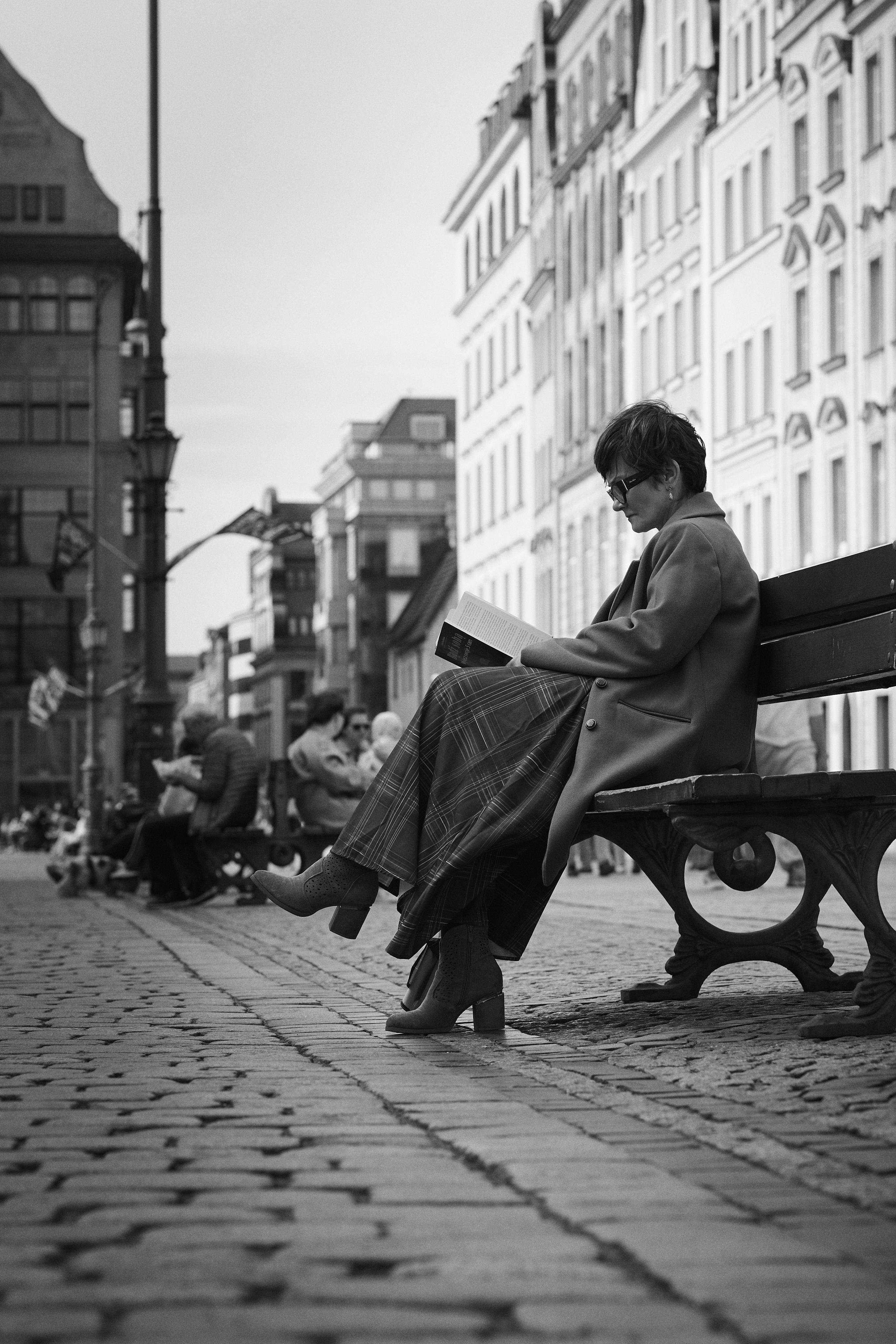 Person wearing sunglasses and a long coat sits on a wooden bench reading a book on a cobblestone street, with other seated pedestrians and tall buildings lining the background in a black-and-white photo.