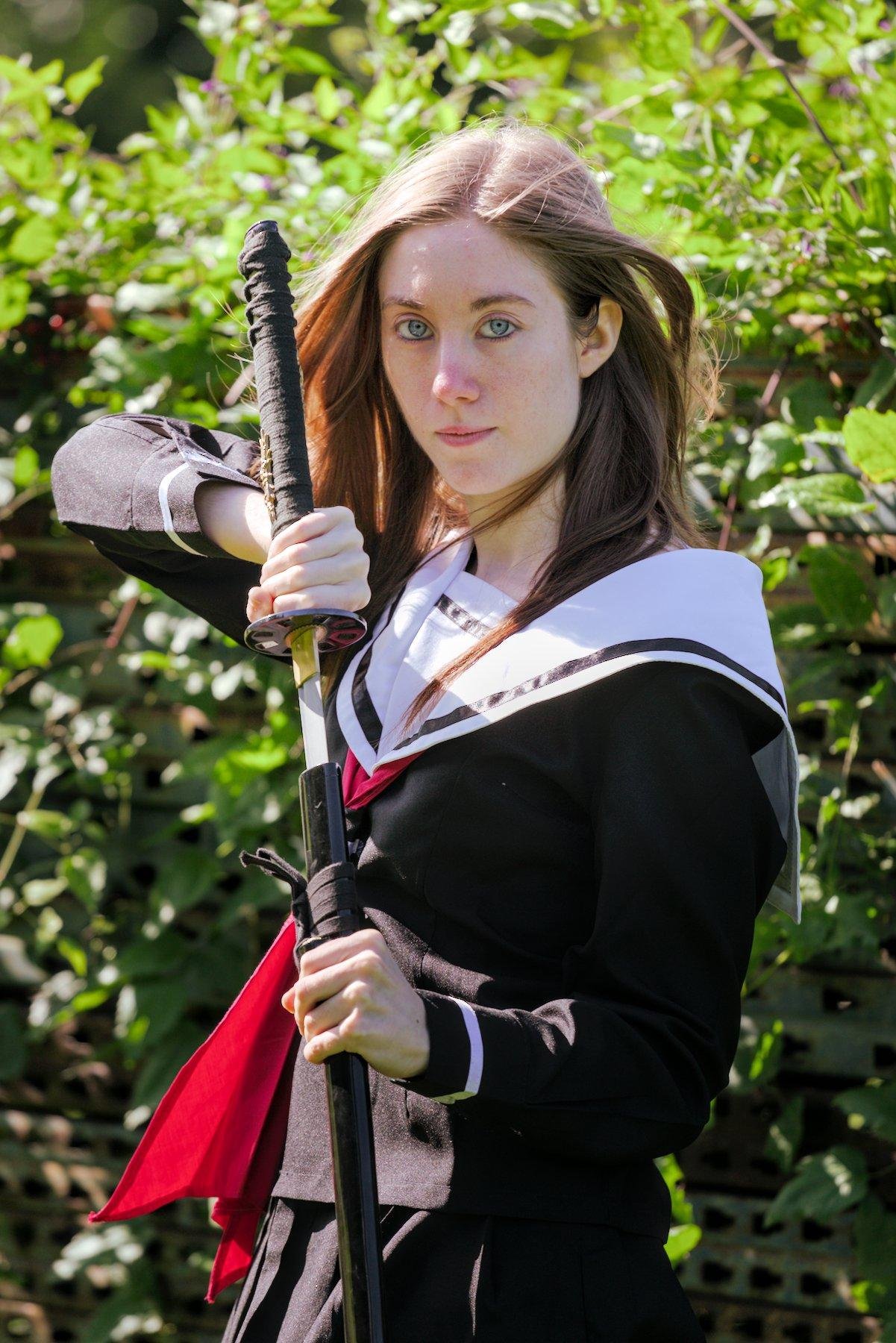 Photo verticale. Une jeune femme rousse en tenue d’écolière japonaise rengaine un katana dans son fourreau, verticalement, en regardant devant elle vers la caméra d’un air déterminé. Le vent fait légèrement voler ses cheveux. Derrière, une haie végétale toute verte