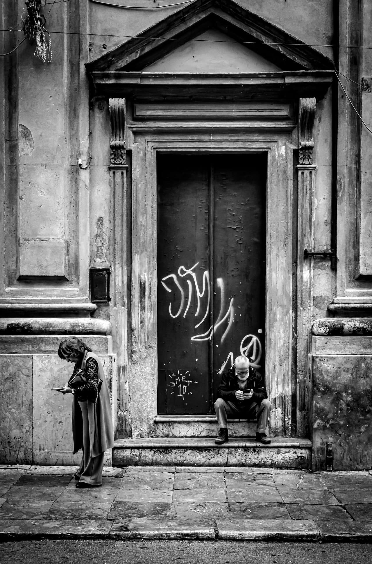 Black-and-white street photo of an old stone building facade with a tall, recessed doorway framed by decorative molding and a triangular pediment; the dark door is covered in graffiti. A woman stands on the left looking down at a phone or small object, while a man sits on the doorstep on the right, also looking at a phone; cobblestone pavement fills the foreground.