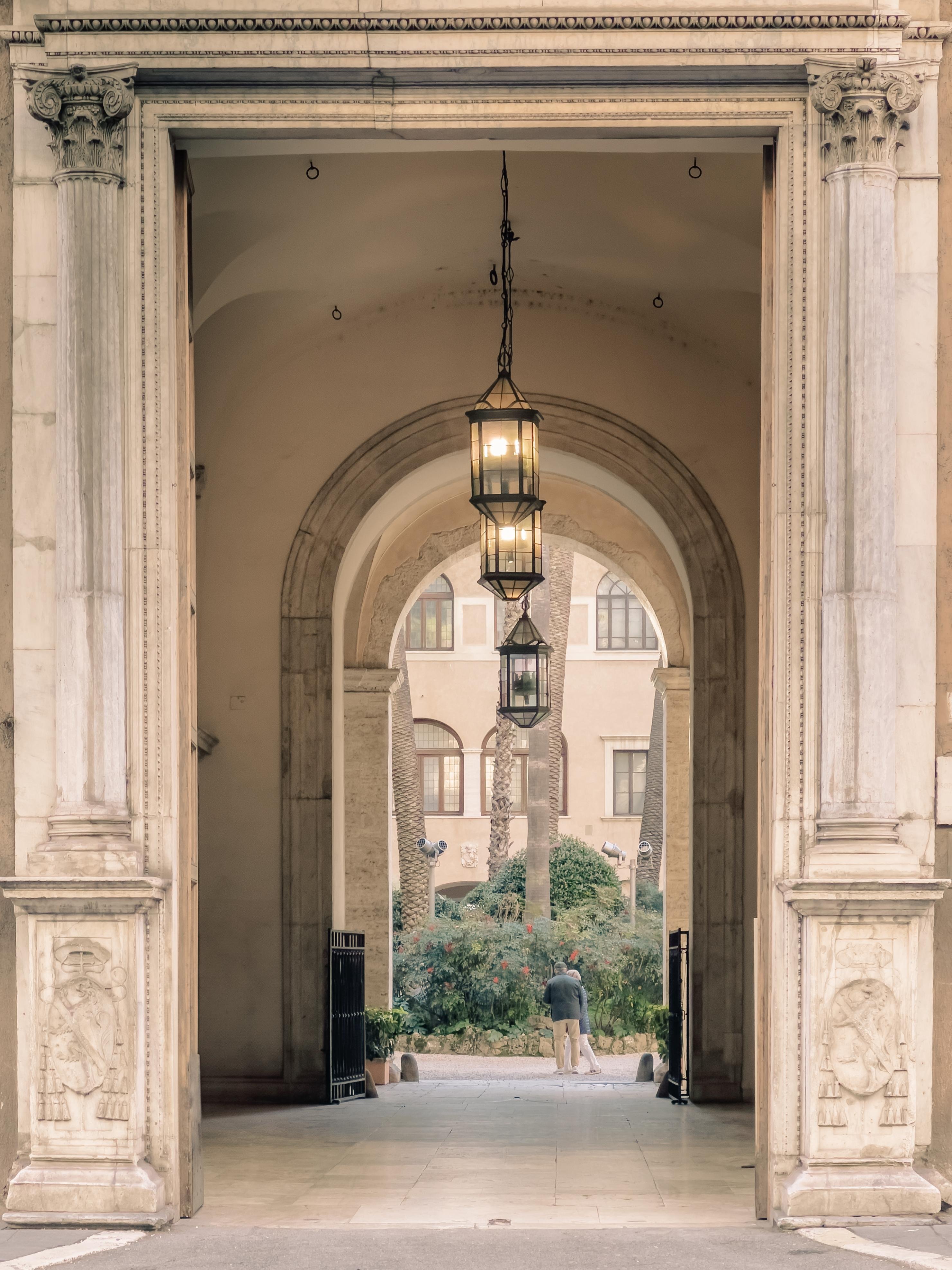 Grande ingresso architettonico con colonne e decorazioni ornate. Nella volta si vedono lanterne appese. Oltre l'ingresso, si può vedere un cortile con piante e una persona che cammina. L'architettura suggerisce uno stile storico o classico.