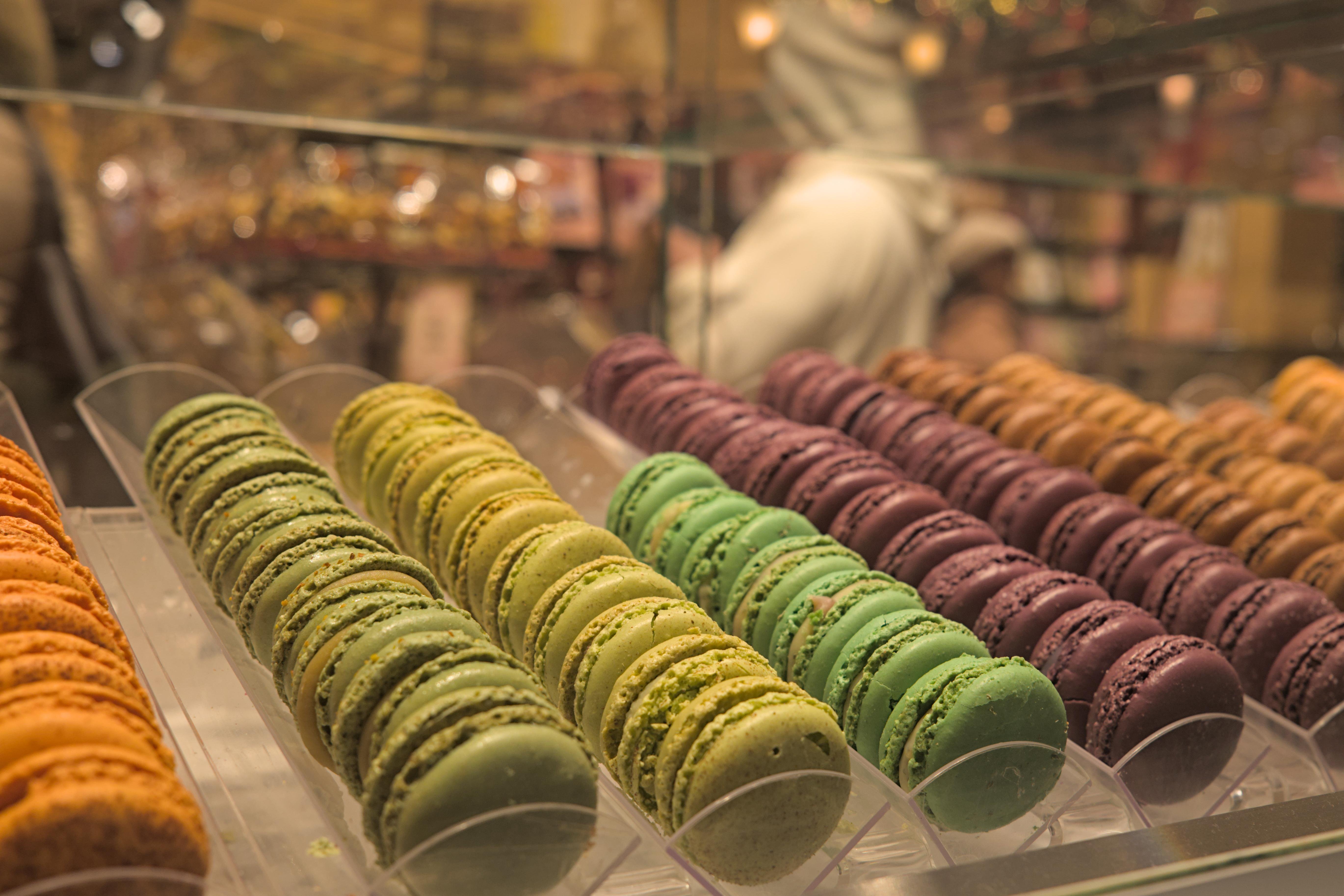 The image shows rows of colorful macarons displayed in a bakery or pastry shop. They are arranged in trays within a glass case.