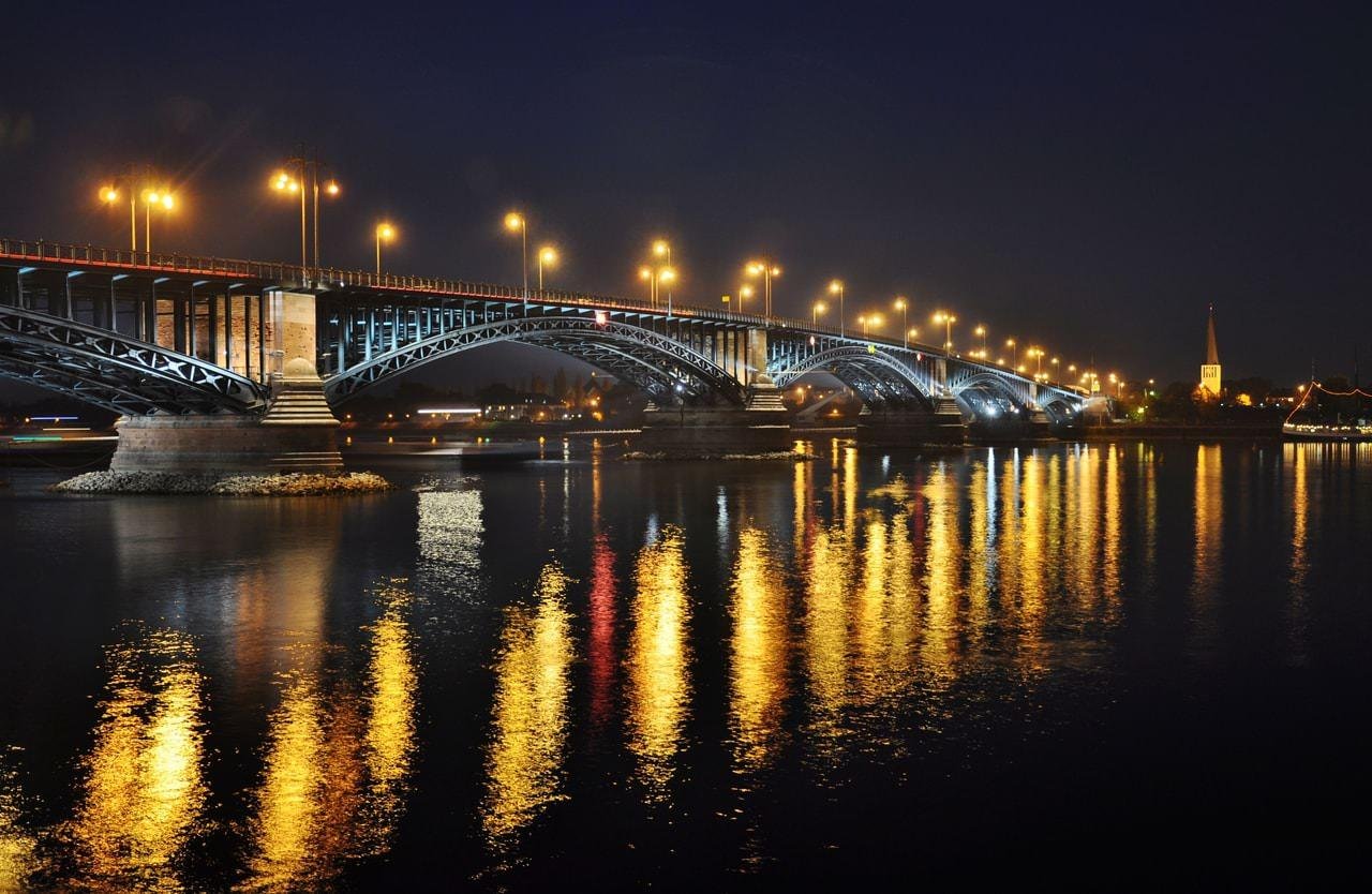 Arched steel bridge at night lit by rows of streetlights, spanning a river with long golden reflections on the water; a church spire is visible on the far right shoreline.