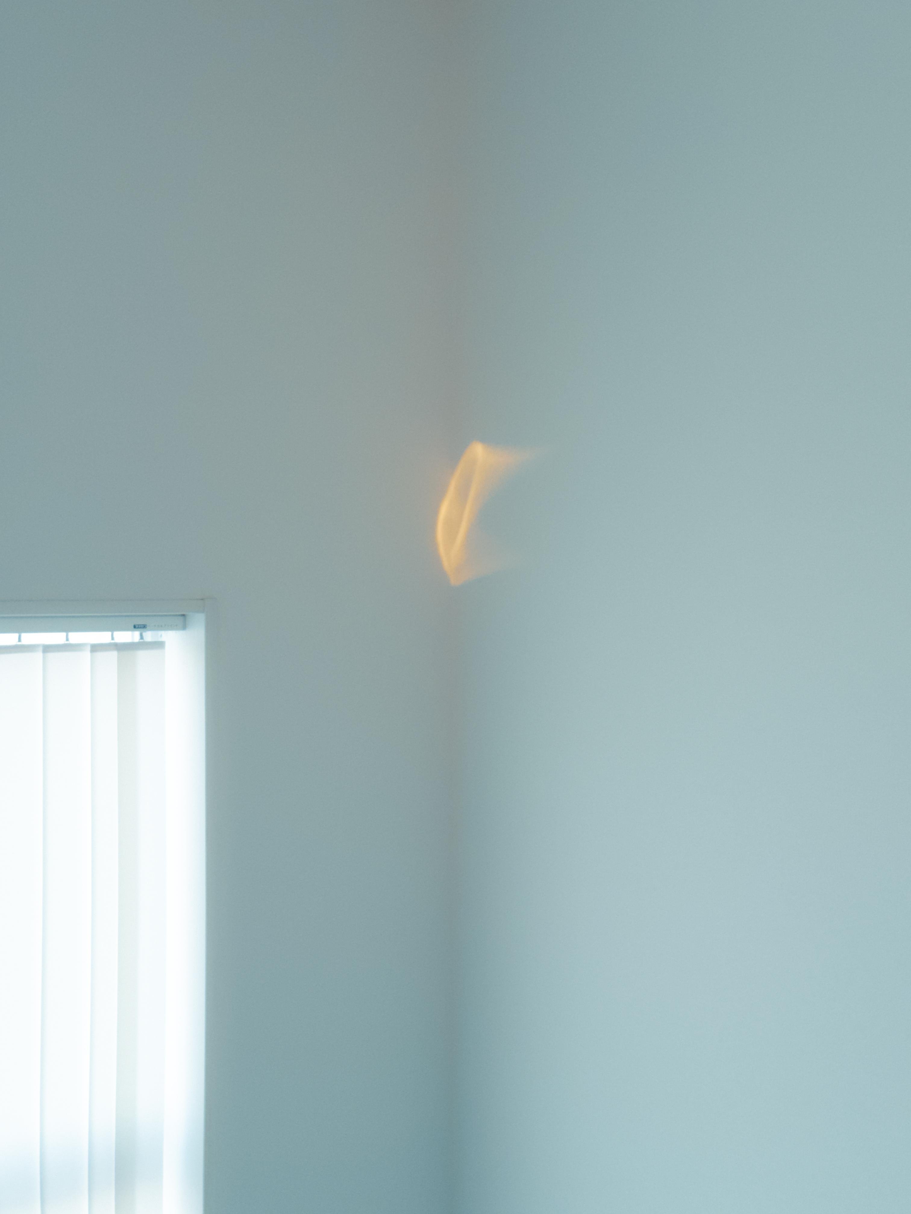 Walls meet in a corner of a room with a soft, warm light reflection. A window with vertical blinds is partially visible on the left side, casting subtle shadows. The overall color scheme is light blue and white, creating a minimalist atmosphere.