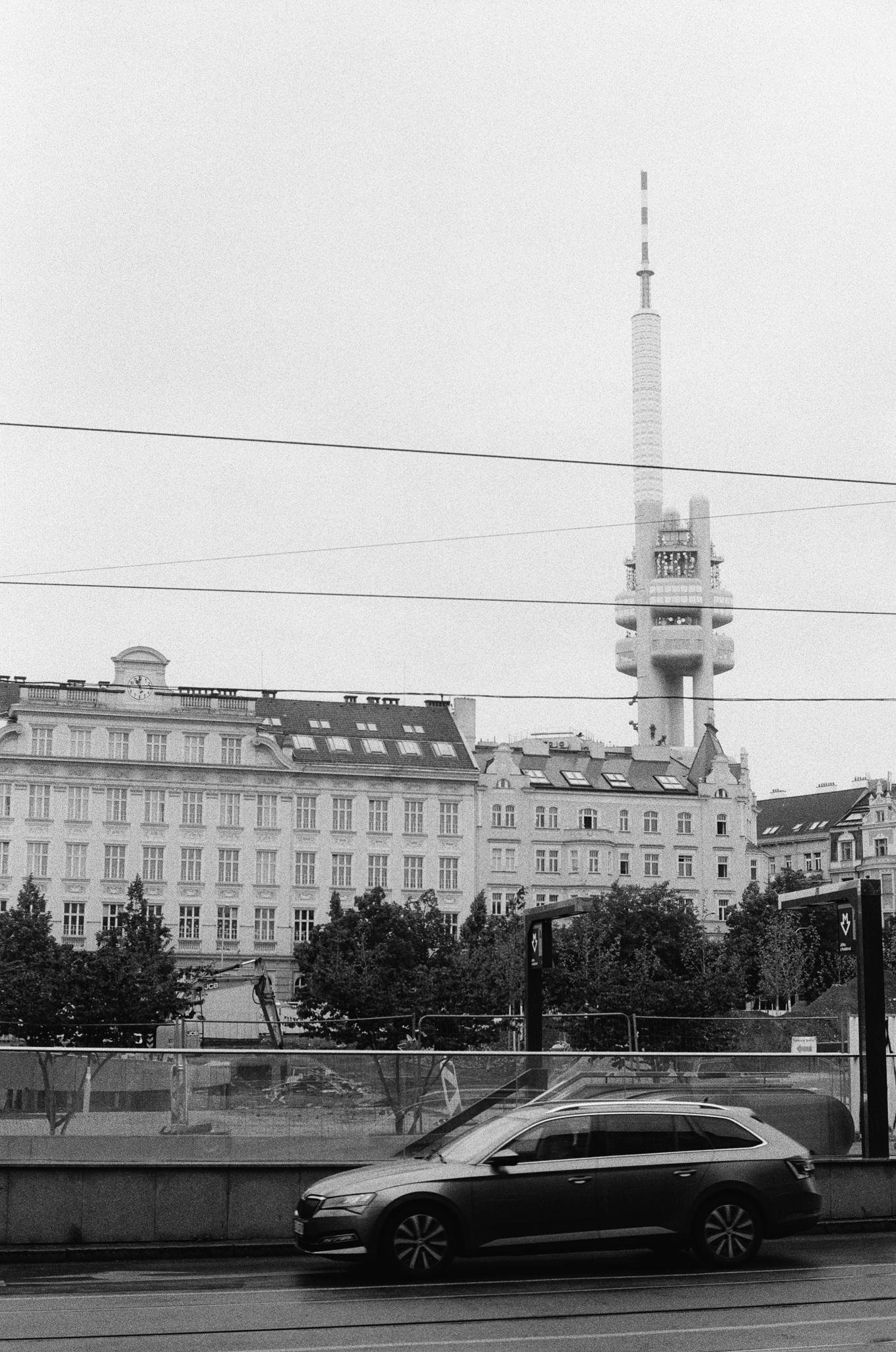 Black-and-white street view with a station wagon parked along a road in the foreground, tram wires crossing the sky, mid-rise historic buildings behind trees, and a tall communications tower with a cylindrical shaft and observation platform rising in the background.