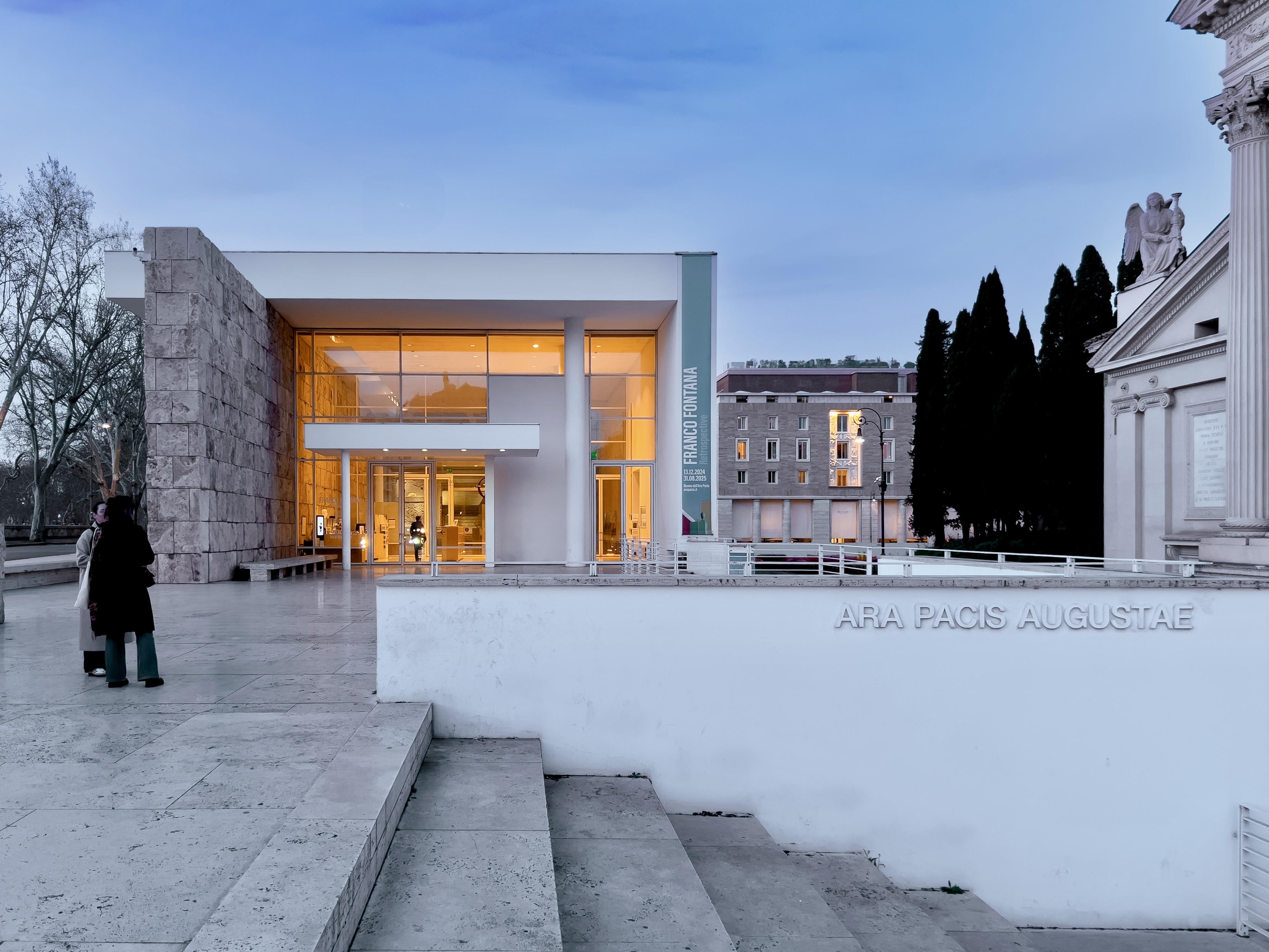 Vista esterna del Museo dell'Ara Pacis a Roma, progettato da Richard Meier. L'edificio moderno, con ampie vetrate e pareti bianche, contrasta con la struttura antica adiacente e la scalinata in travertino. In primo piano, una scritta incisa recita "ARA PACIS AUGUSTAE". Il cielo è crepuscolare.