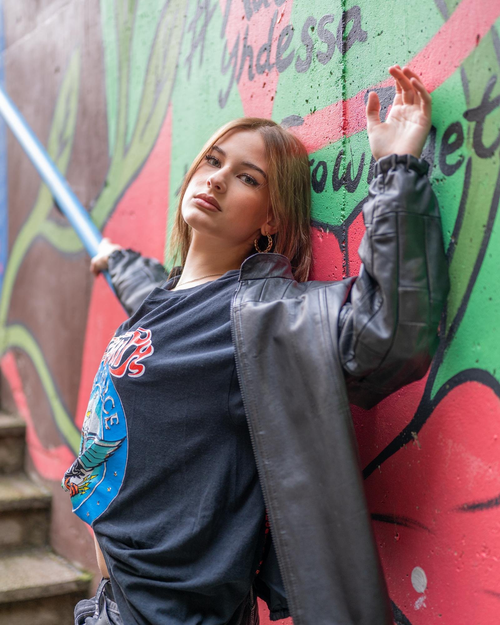 Young person leaning against a mural-painted wall, wearing a black leather jacket and graphic t-shirt with a design featuring an eagle and the word "peace." The wall behind is green and red with partially visible text. Their arm is raised, and they are holding a blue object, possibly a light saber. Steps are visible to the side.