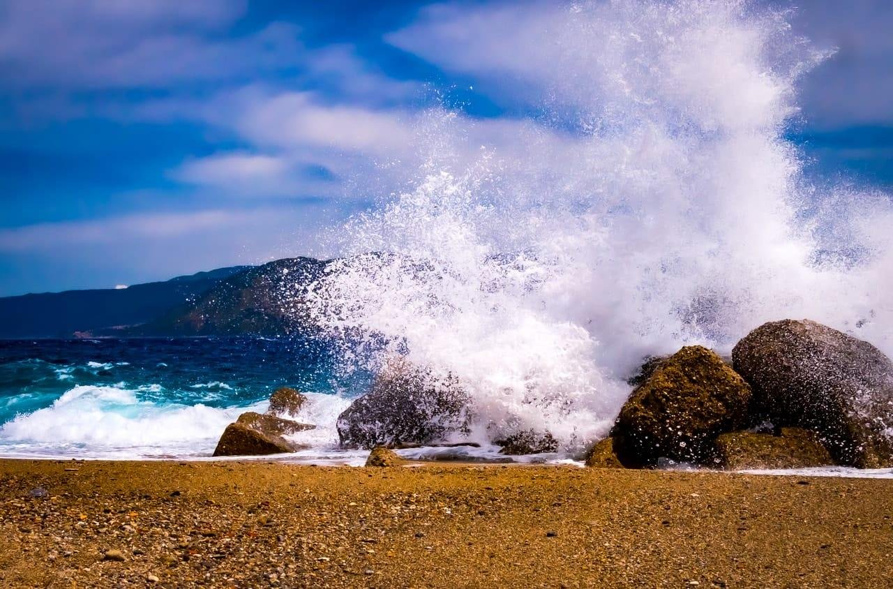 Large ocean wave crashing into dark boulders on a sandy beach, sending white spray high into the air; blue water and a low, dark coastline under a partly cloudy sky in the background.