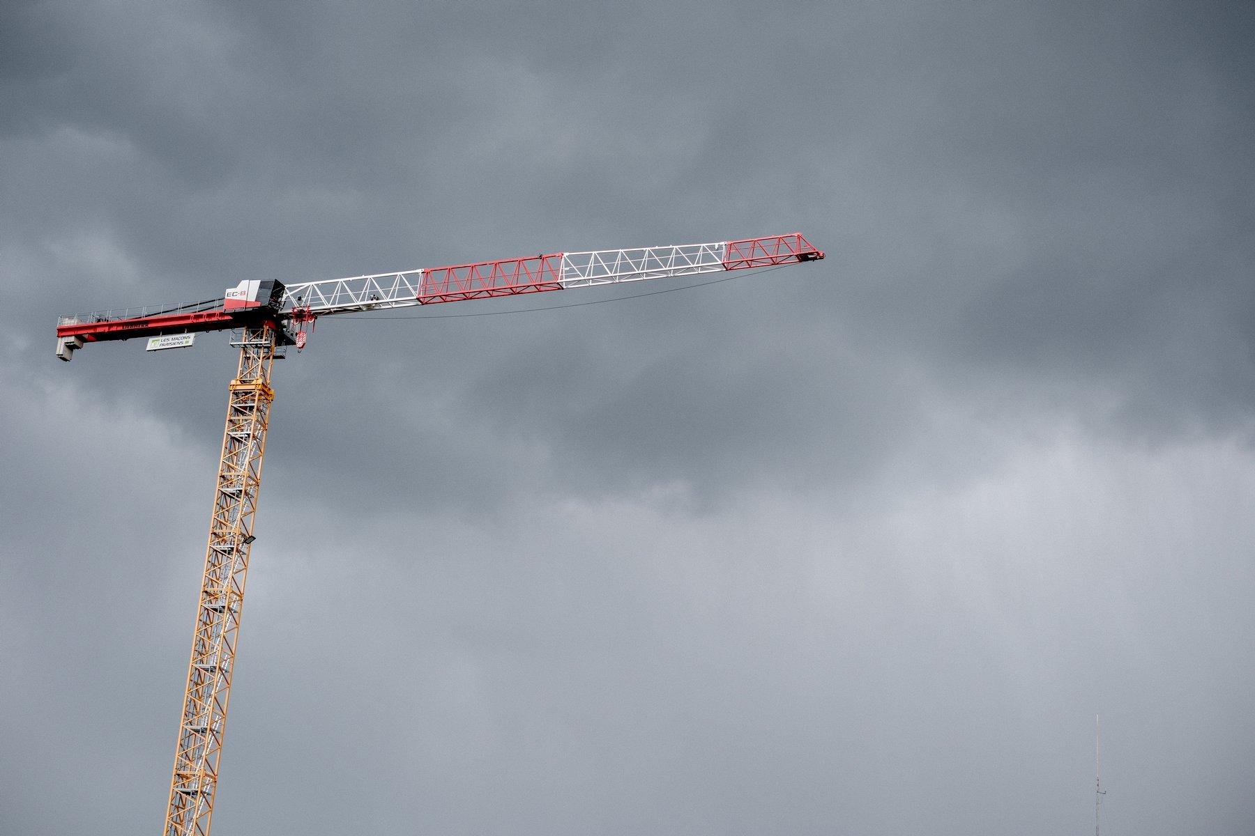 Photo horizontale. Une grue jaune sur la partie verticale, rouge et blanche sur la partie horizontale, sur fond de ciel gris bicolore, foncé en haut de l’image et plus clair sur le tiers bas. On devine un rideau de pluie sur la limite des 2 gris