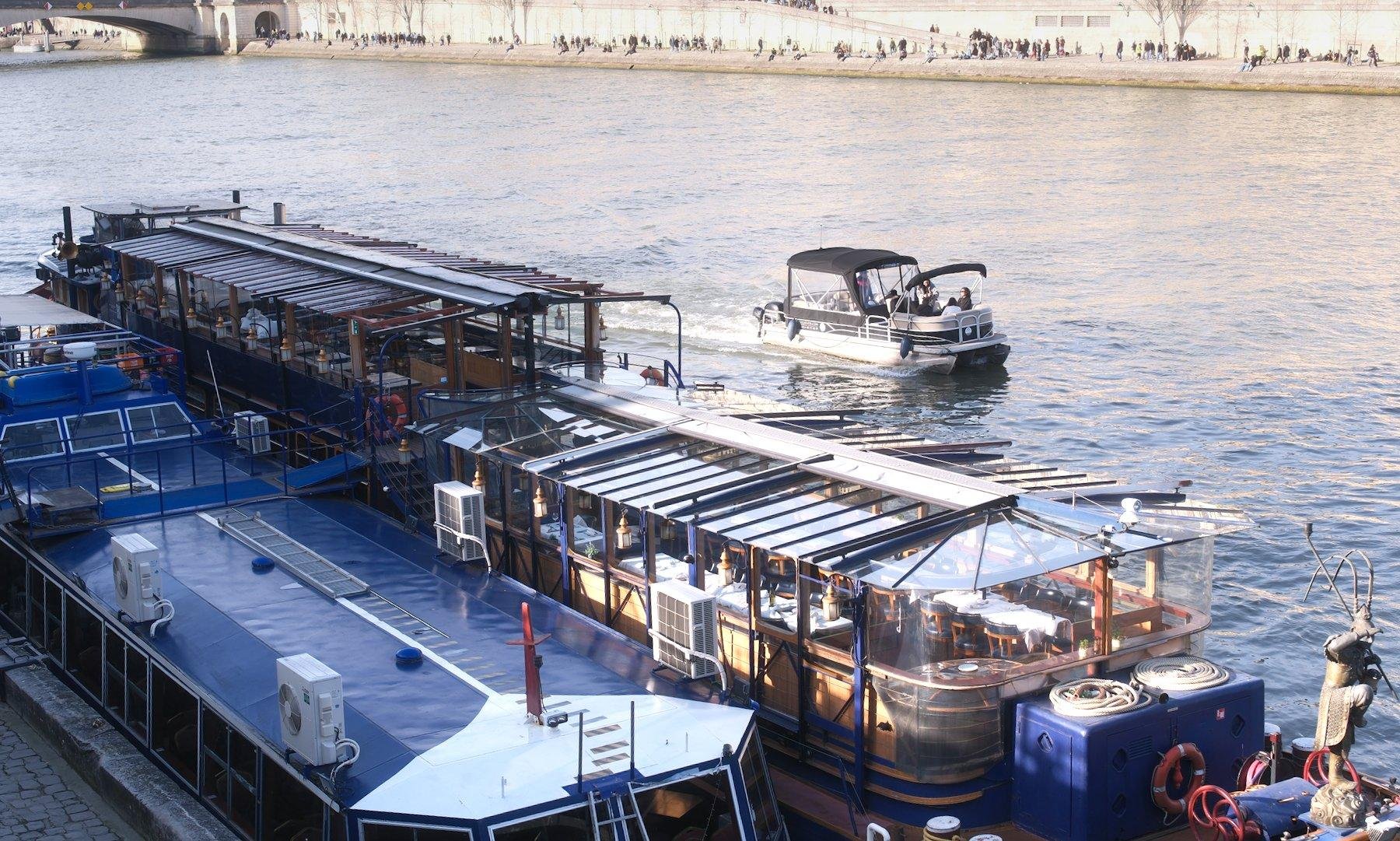 Photo horizontale. Vue sur la Seine où 2 péniches sont amarrées côte à côte, et tout petit bateau passe, leur taille contrastant beaucoup