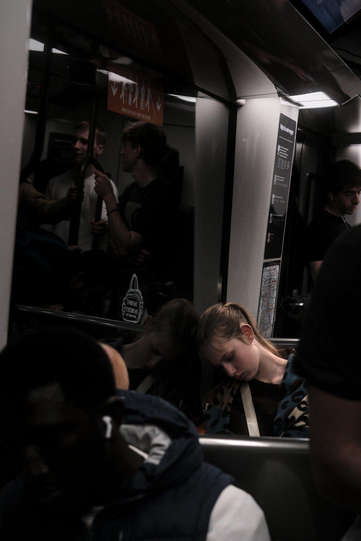 Photo verticale. Dans une rame de métro, une jeune femme assise dans un carré de 4 a sa tête posée contre la vitre, regardant son téléphone plus bas, l’air de s’ennuyer ferme. Son reflet dans la vitre double la dose d’ennuie