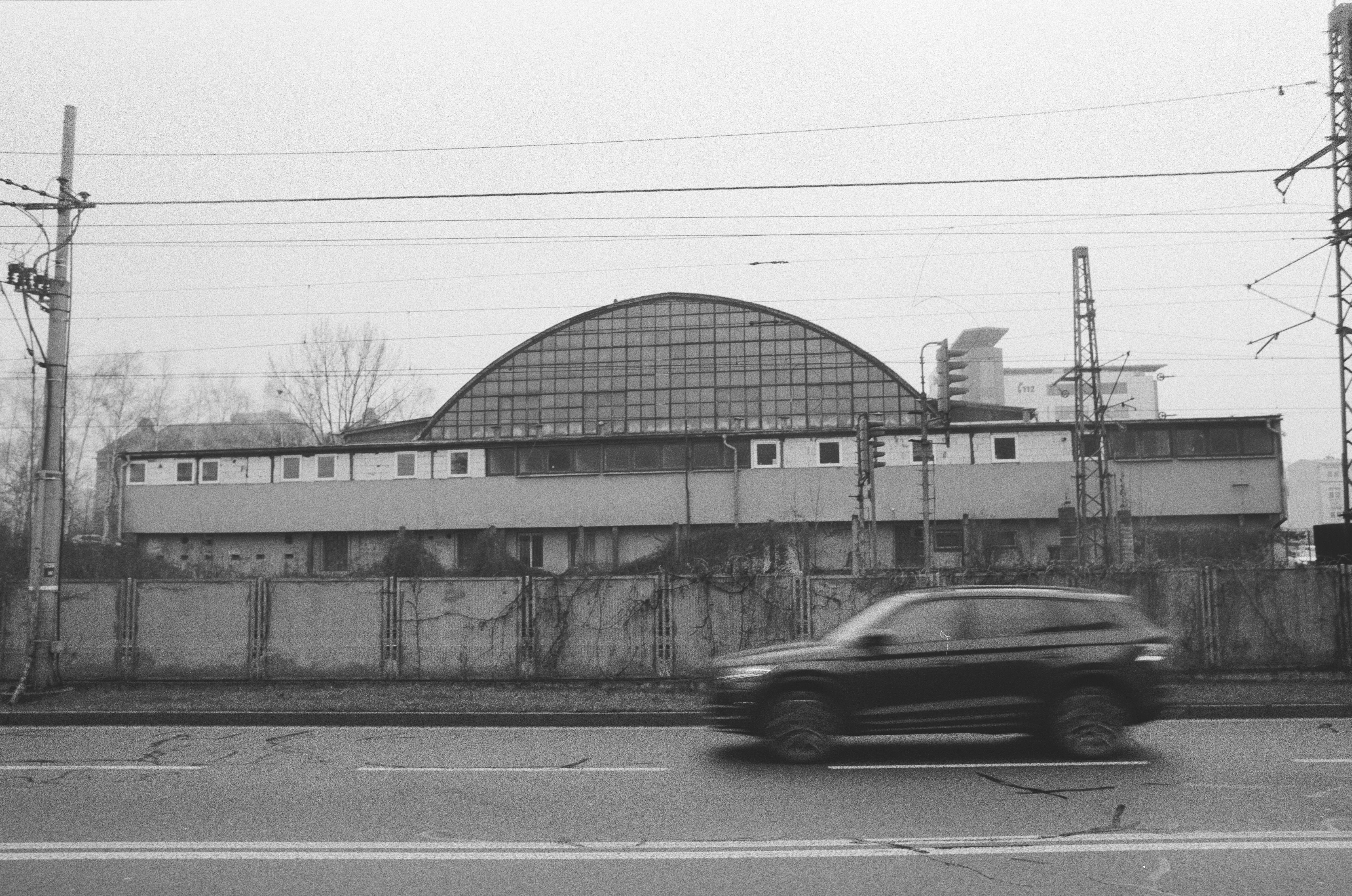 A monochrome photo of a delapitated building with concrete wall separating it from diagonal road with motion blurred car on it.
