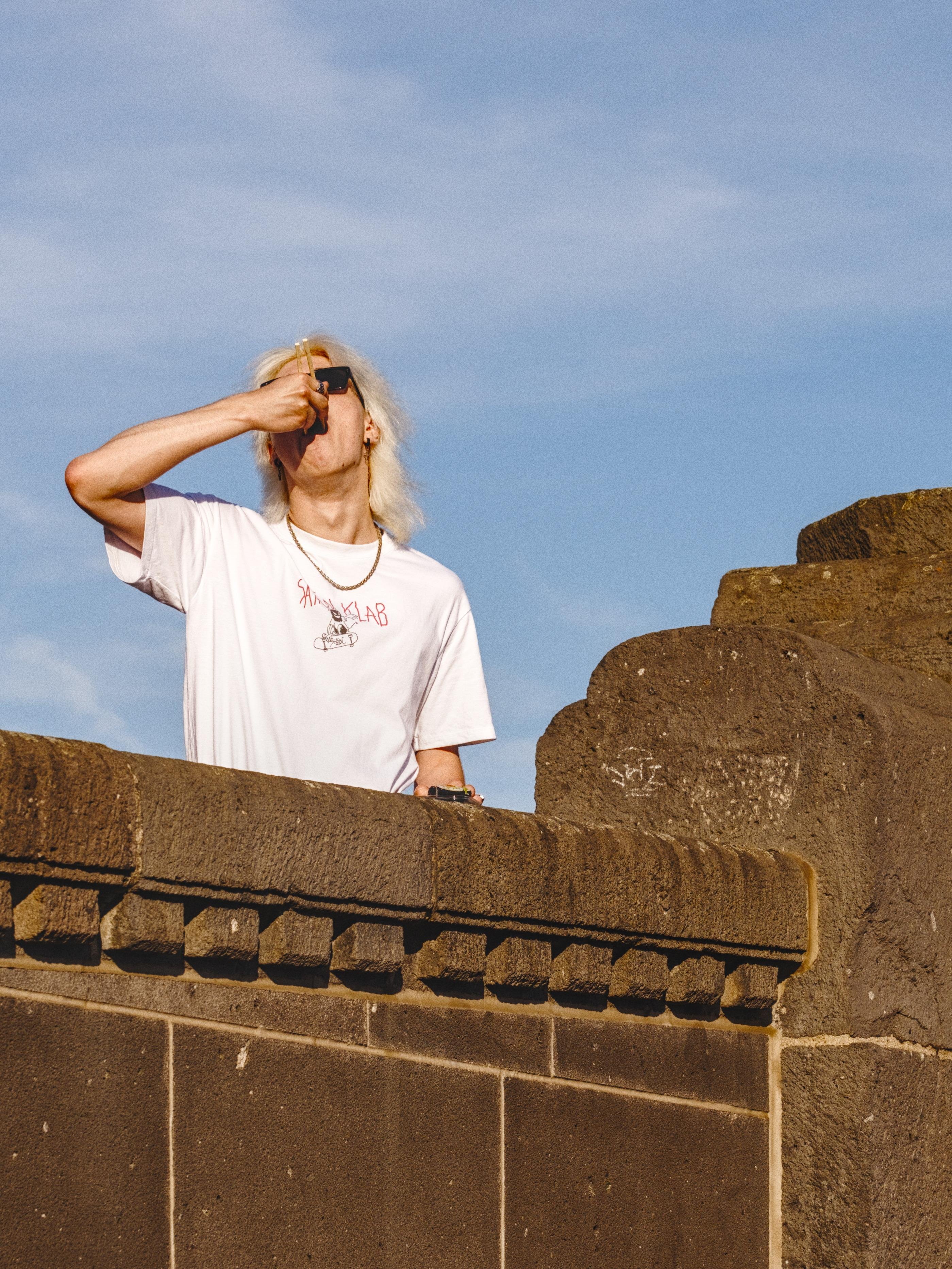 A man eating sushi by plunging his sticks from above his head, in an almost exaggerated gesture. He's wearing a white designer shirt with a necklace and is standing atop a bridge, of which only the side is visible. 