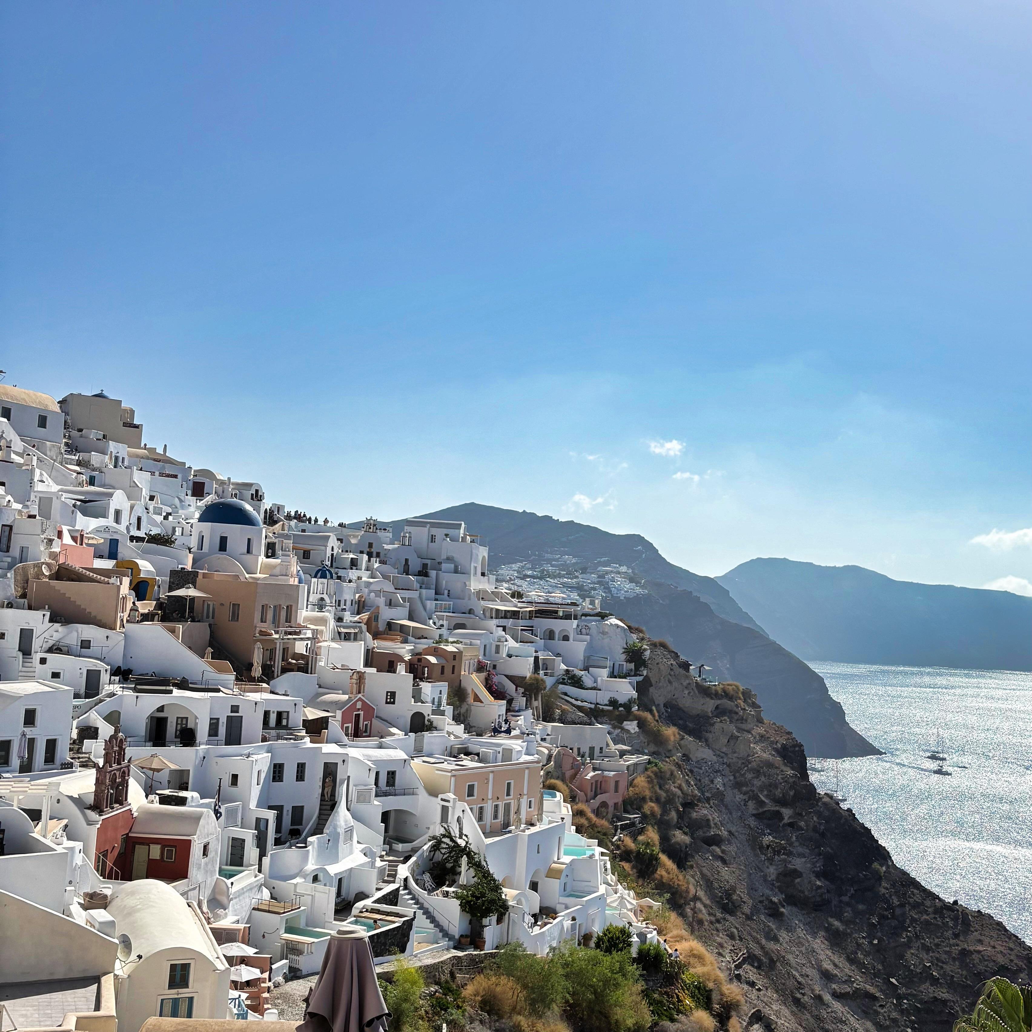 White and pastel-colored buildings with domed roofs cascading down a steep hillside on the left. A clear blue sky and a sunlit sea with a sailboat in the distance on the right. Rugged cliffs extend into the sea, creating a dramatic coastal landscape.
