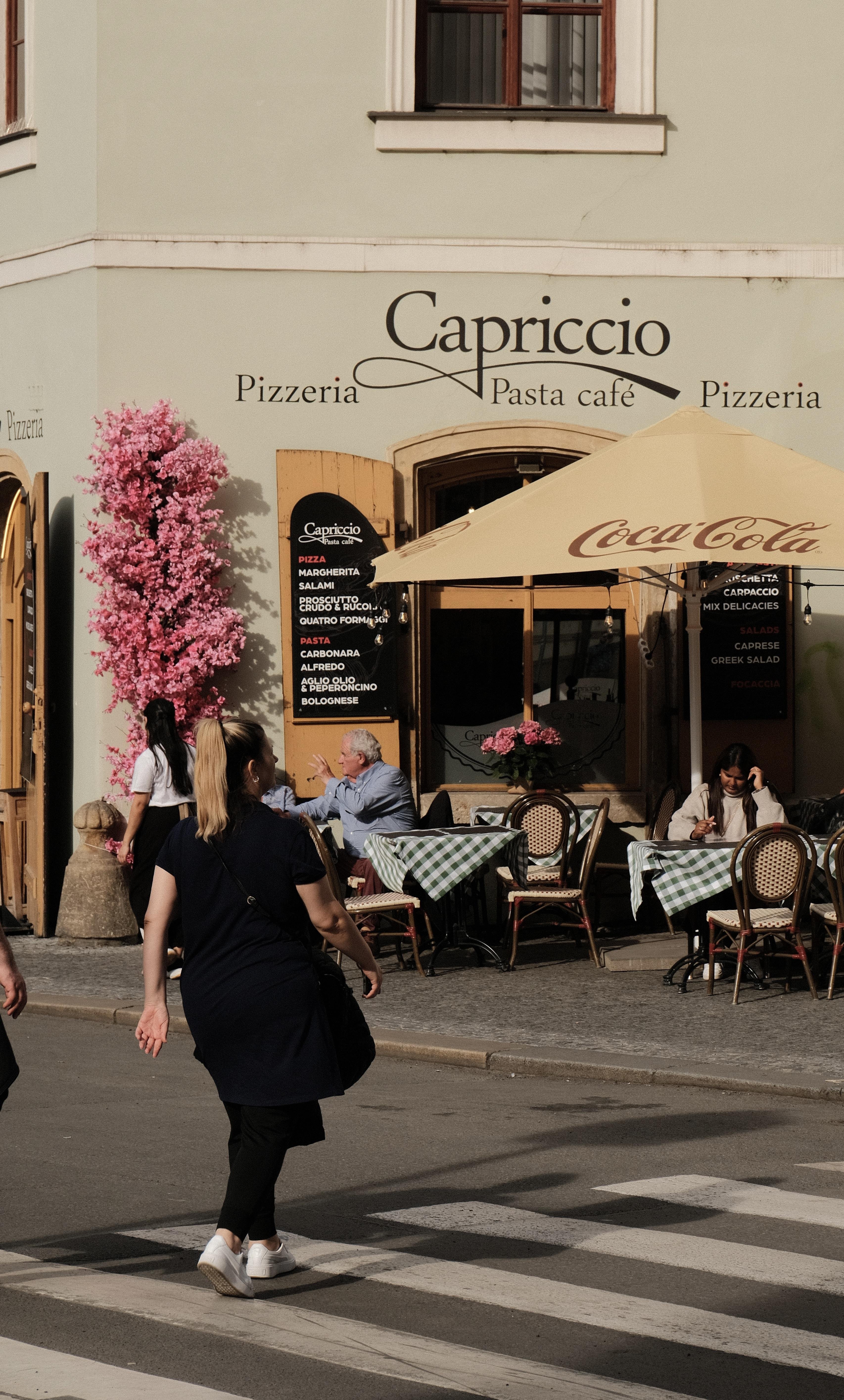 Street view of a Café called Capriccio with outdoor tables under a Cream-white Coca‑Cola umbrella; a person crosses a zebra crossing in the foreground, patrons sit and talk near the entrance of the Café, and a tall pink flowering plant stands beside the doorway.