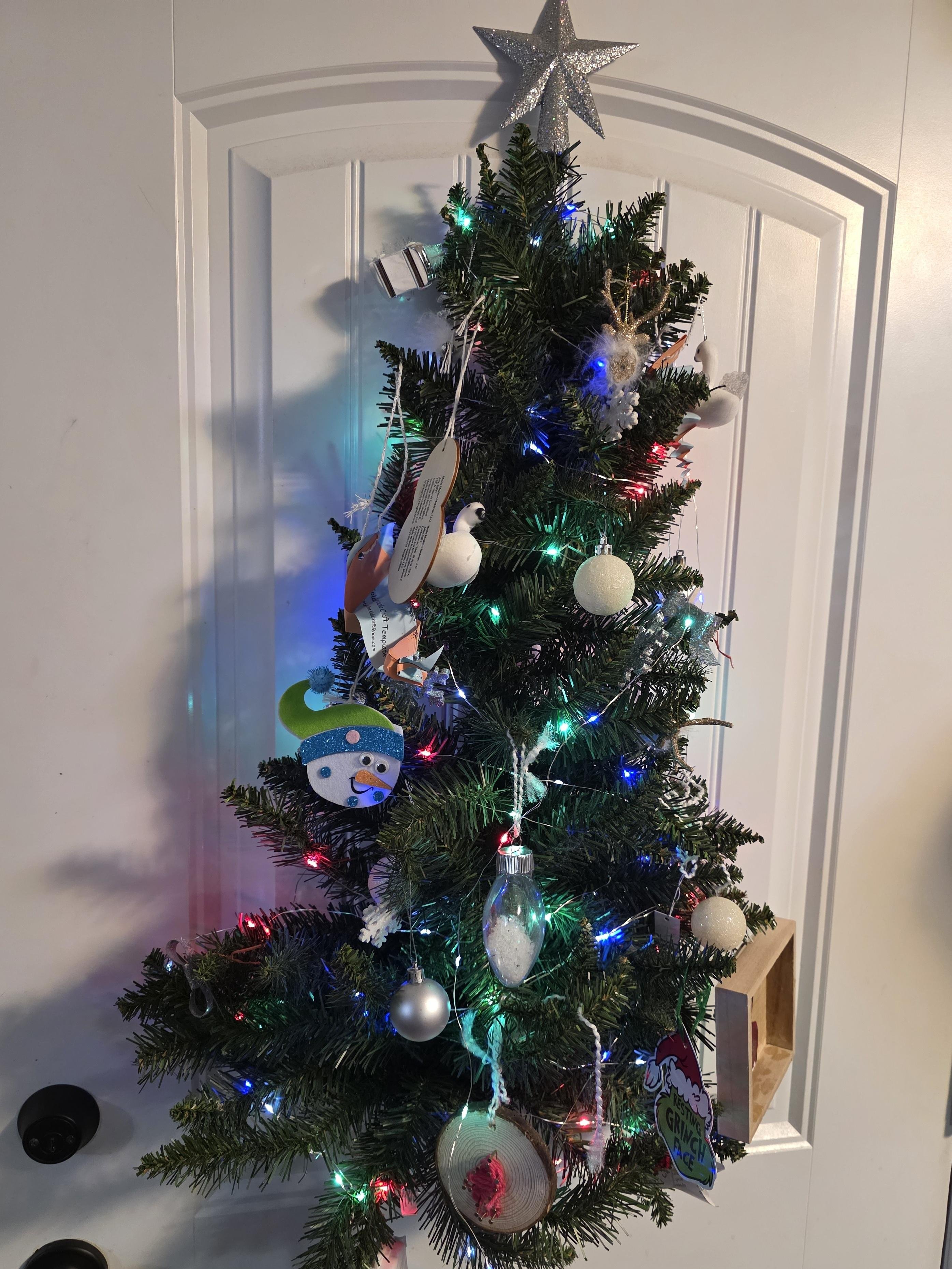 Artificial Christmas tree decorated with colorful string lights and various ornaments, including a snowman, white spheres, and a Grinch illustration. A silver star tops the tree, which stands in front of a white paneled door.