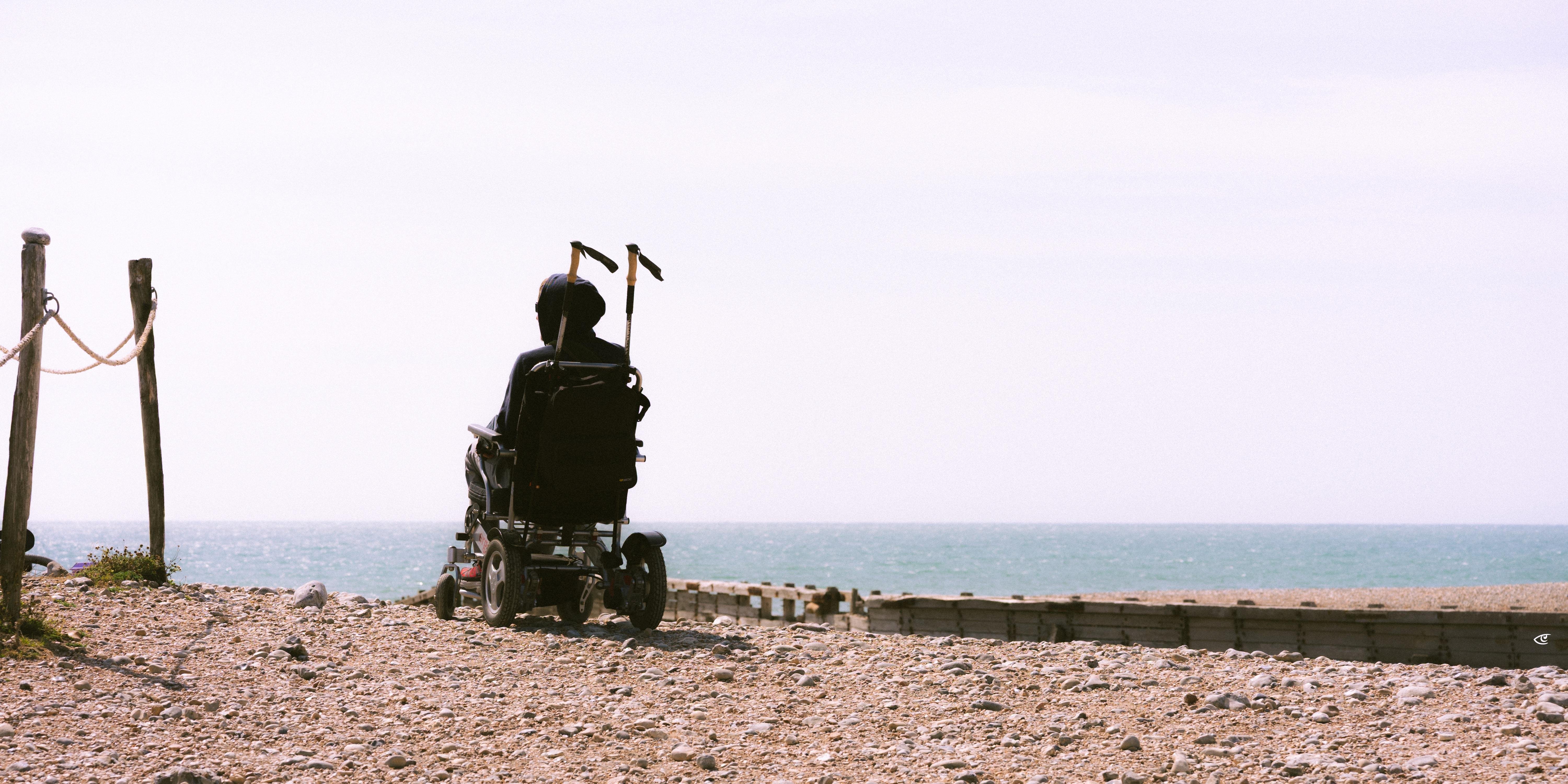 Person in a motorized wheelchair viewed from behind on a rocky beach, facing the ocean; wooden posts with rope on the left and a low seawall along the shoreline under a pale sky.
