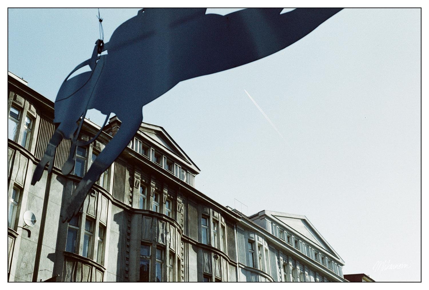 The image shows a street view with a decorative silhouette of a horse or rider suspended in the foreground. In the background, there is a row of historic buildings with decorative facades, and the sky is clear with a visible contrail.