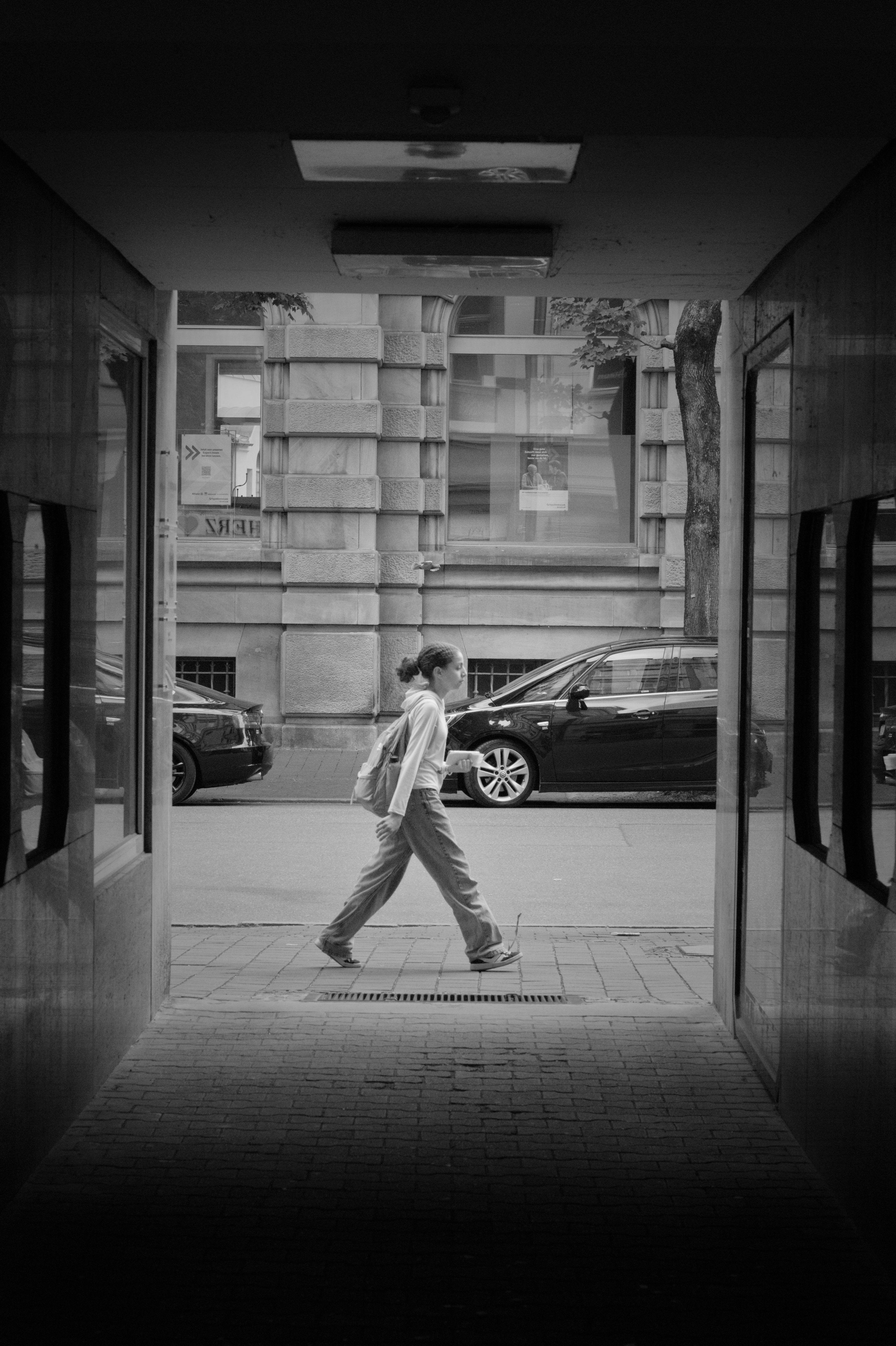 Black-and-white view from inside a shadowed passageway framing a sidewalk and street; a woman with a backpack walks left to right past parked cars, with a stone building facade and a tree in the background.