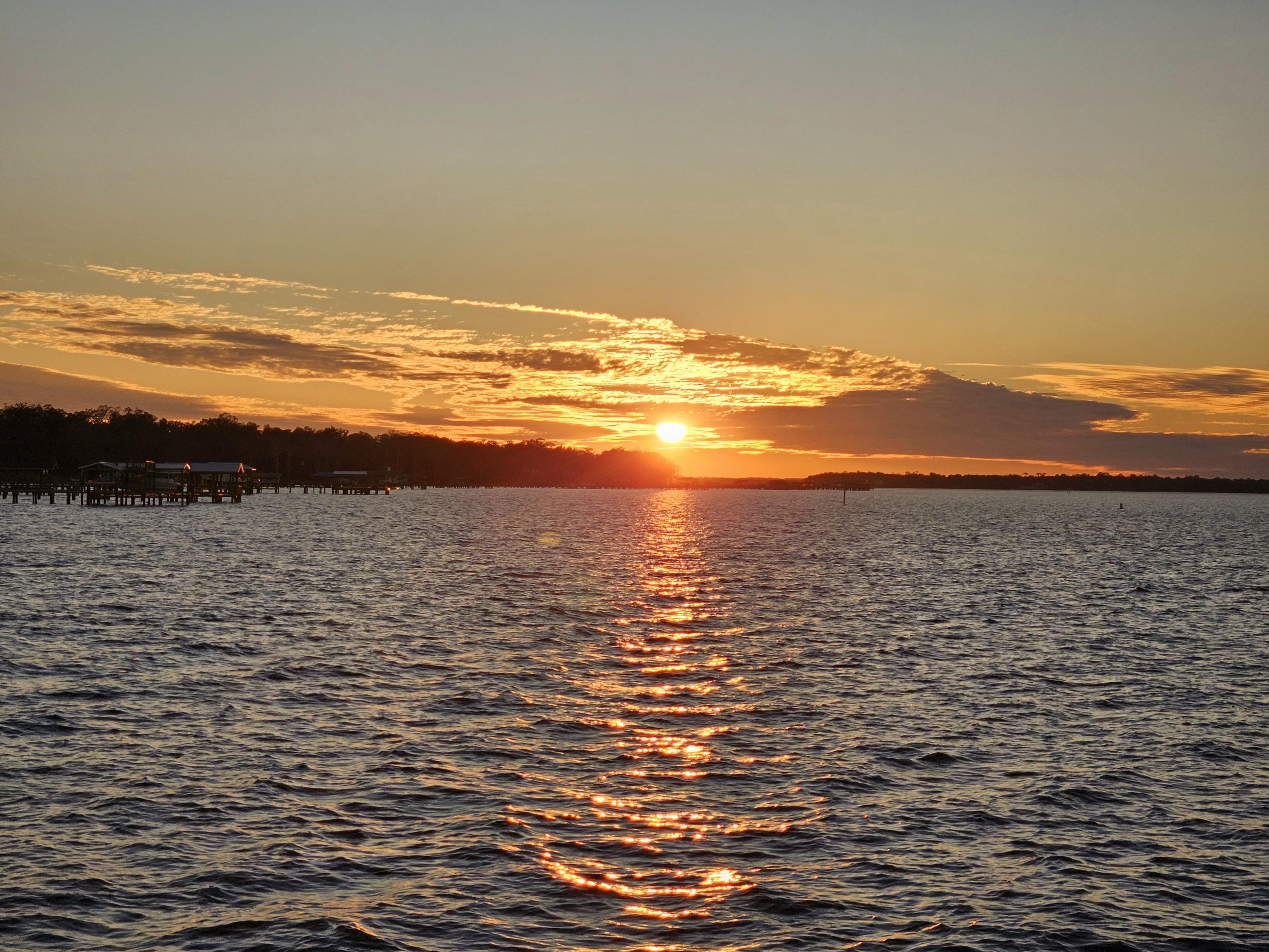 Sun setting over a large body of water, casting a golden reflection across the surface. Clouds partially cover the sky, illuminated with warm hues. Silhouettes of trees and a dock are visible along the horizon.