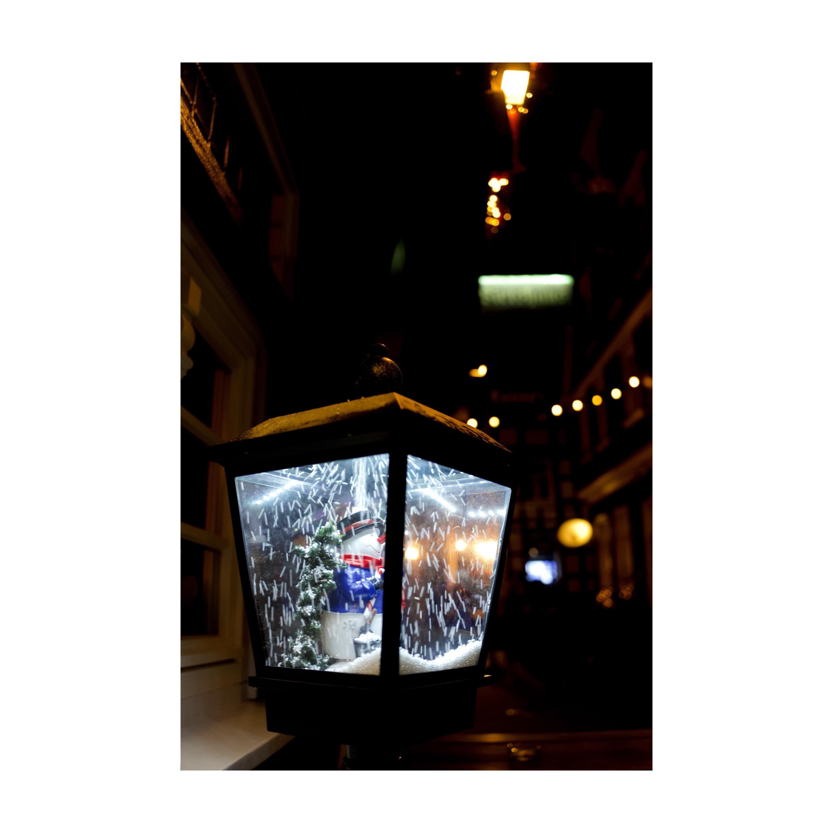 Leuchtende Laterne mit kitschiger Schneekugel-Szene und Lichterkette in nächtlicher Straßenkulisse, stimmungsvoll fotografiert.