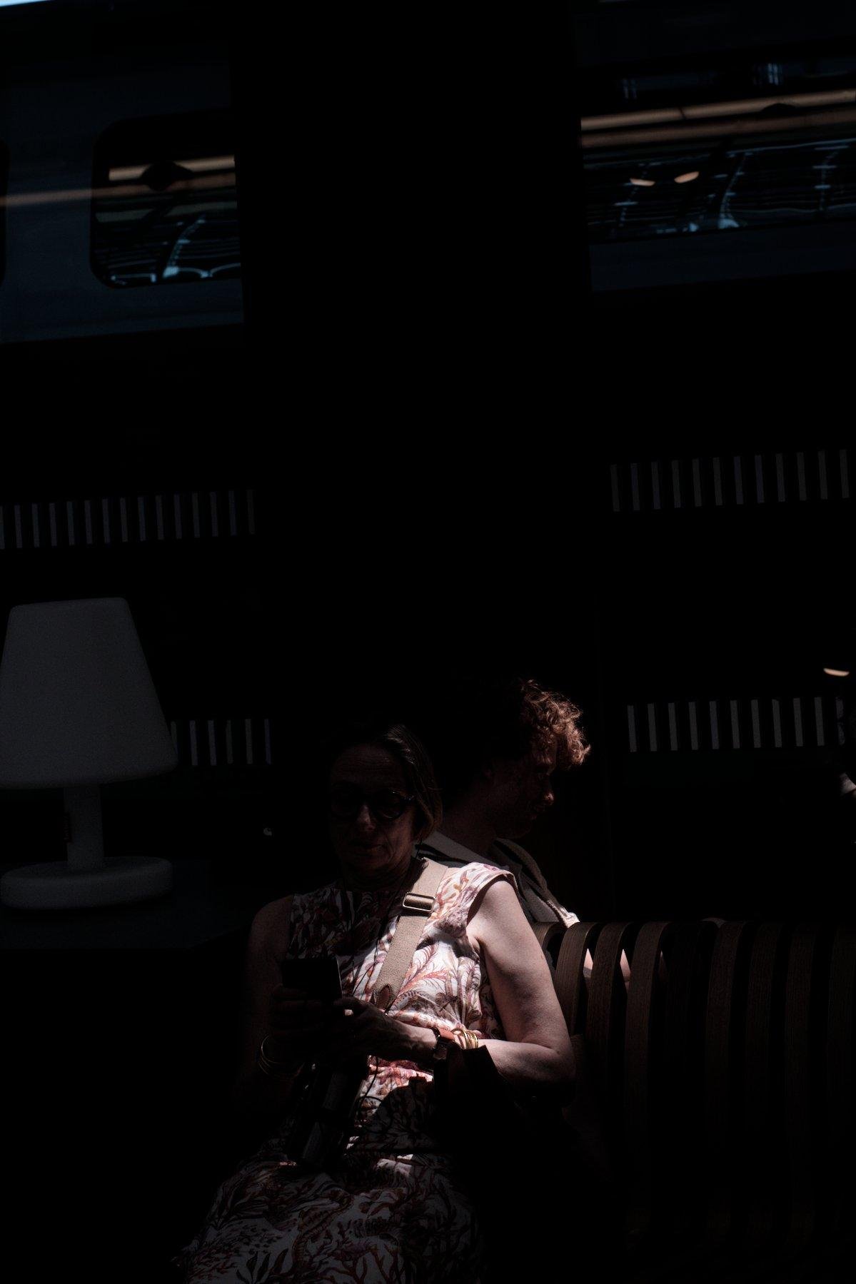 Photo verticale. Dans l’obscurité, un raie de lumière éclaire les cheveux roux bouclés d’un homme assis, dos à dos avec une dame éclairée sur l’épaule et sa robe blanche aux motifs végétaux. En haut, on devine un train à l’arrêt, ils attendent leur train en gare donc.