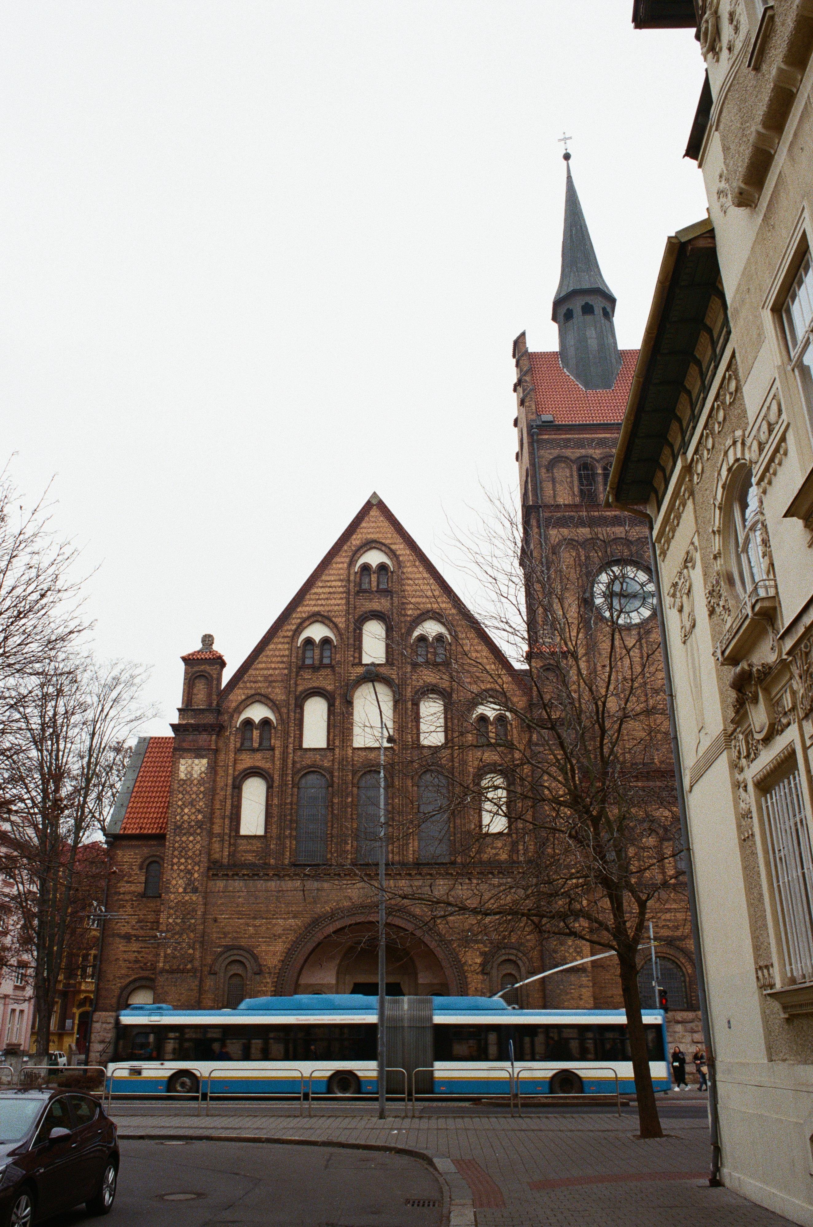 The image depicts a large brick building with a pointed roof and a clock tower, possibly a church or historical building. In front of the building, there is a street with a blue and white trolleybus passing by, along with a few trees and cars. The architecture features arched windows and detailed masonry.