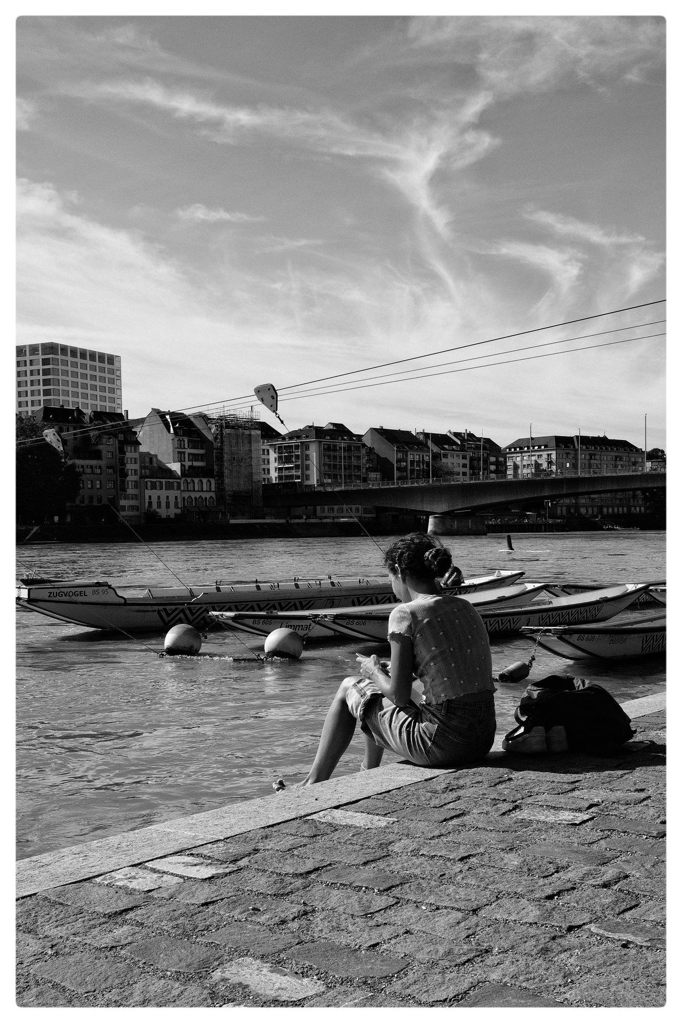 A person sits on a stone embankment with water in the foreground. Several rowboats are docked nearby. A bridge spans the river, leading to a row of buildings. Sparse, wispy clouds fill the sky. The scene is captured in black and white.