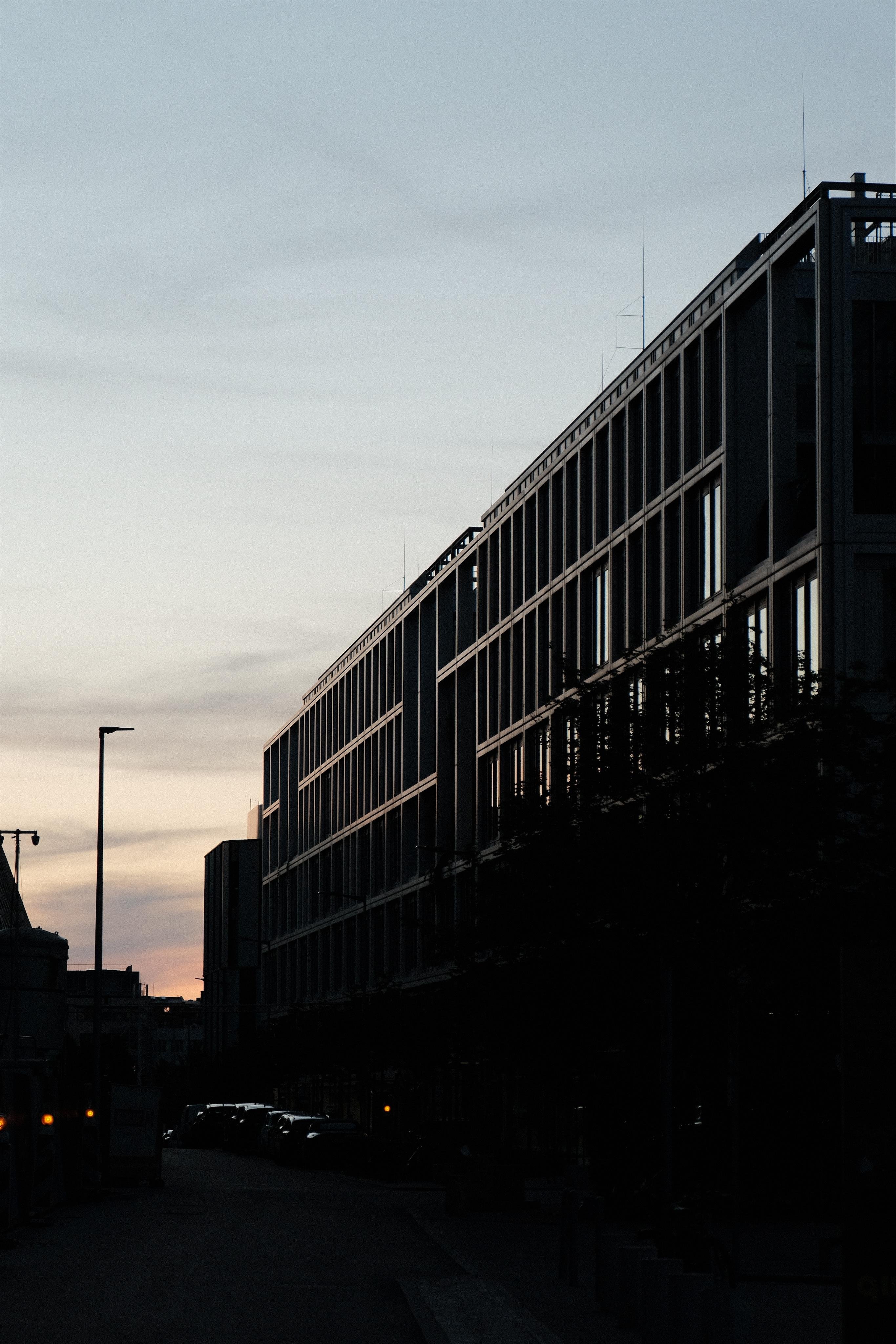 View of a long, multi-story building with a grid of windows on the right, silhouetted against a dusk sky; a street with parked cars and a single streetlight runs along the bottom left, however this part of the shot is already quite dark.