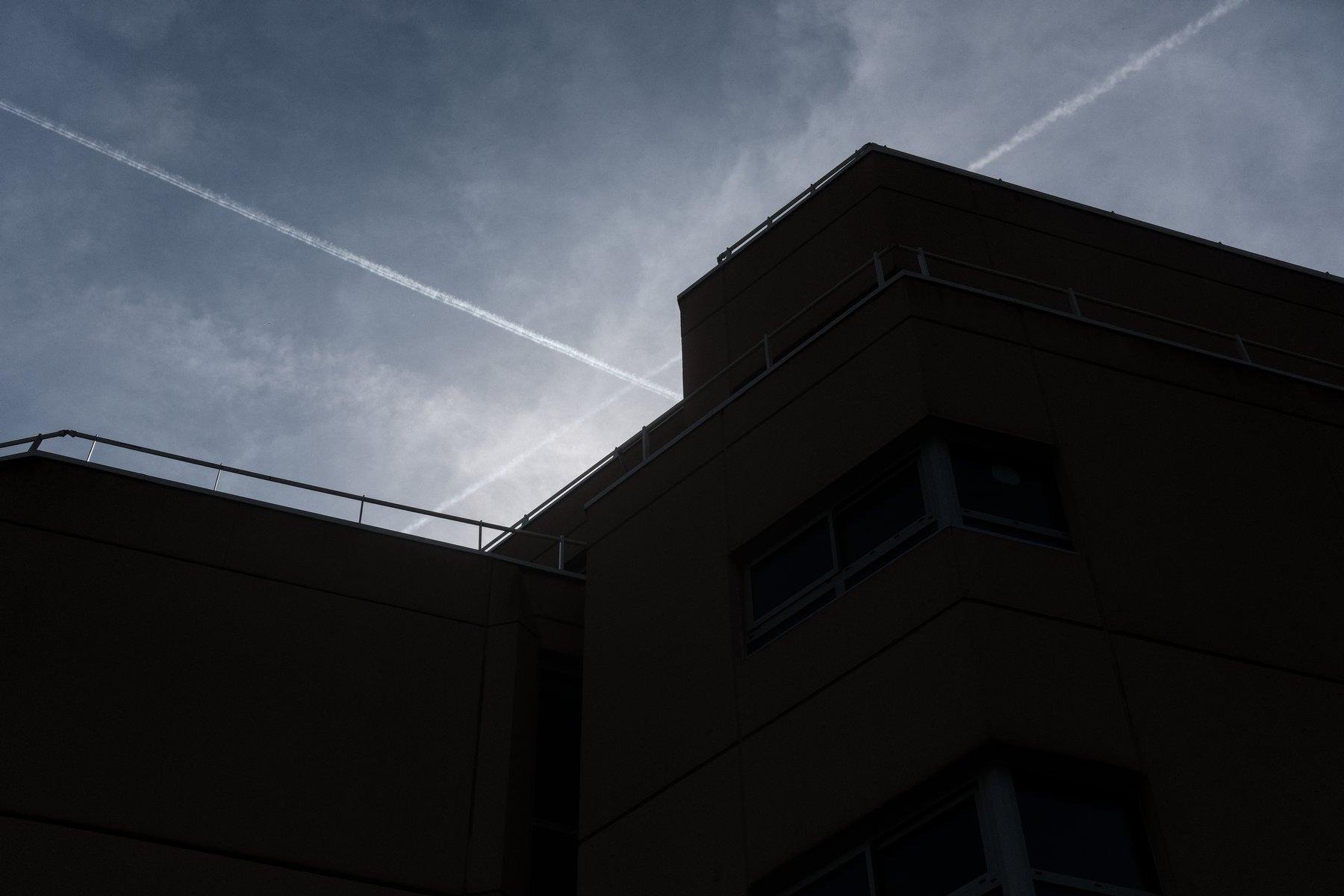 Photo horizontale. Contre-plongée sur un bâtiment très anguleux à contre jour sur un ciel bleu marqué du passage de 2 avions et de volutes de nuages