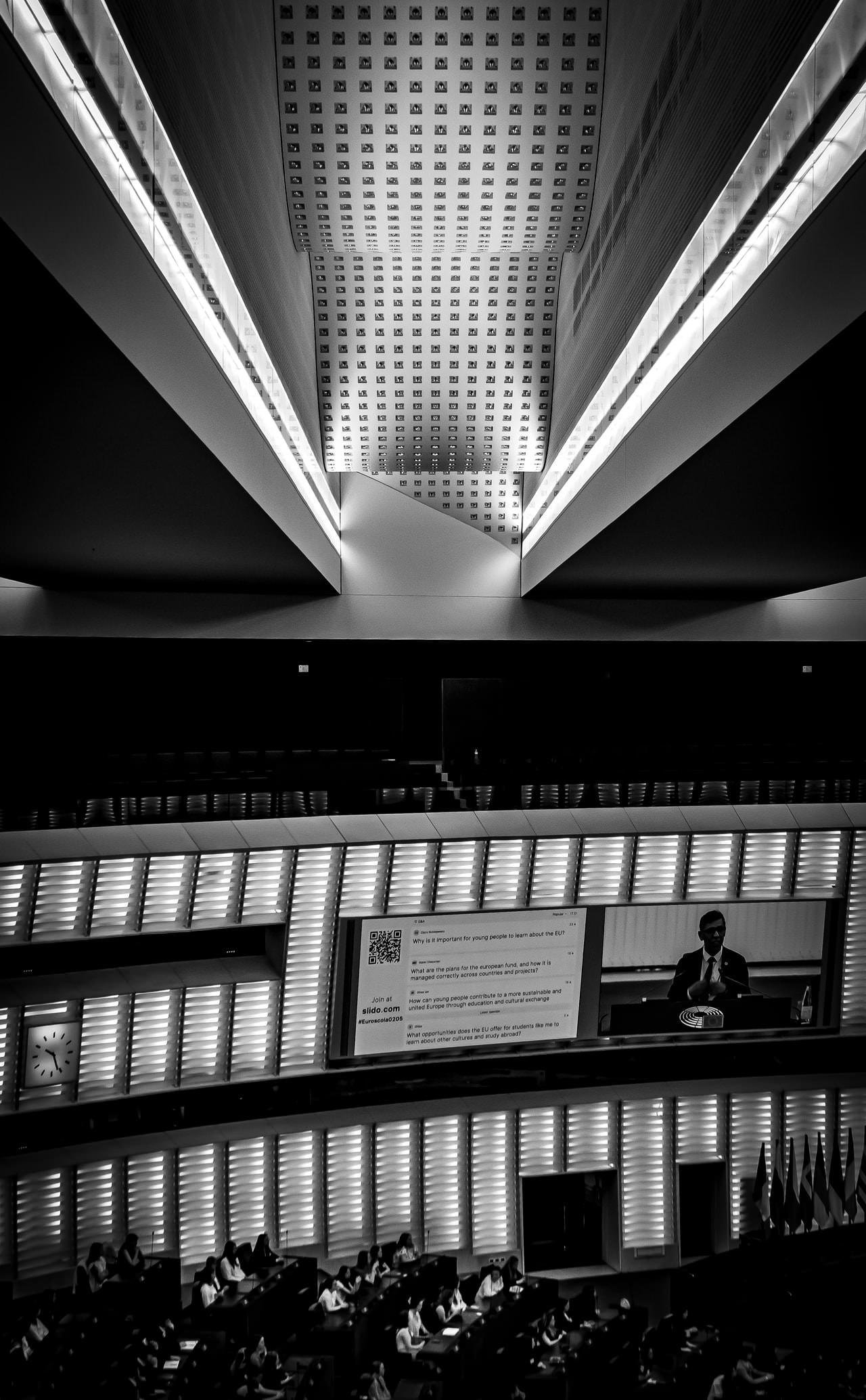 Black and white image of a modern conference hall with an illuminated geometric ceiling pattern. The lower section features an audience seated in rows, a large display screen showing text and QR codes, and a speaker at a podium. Walls are lined with vertical lights, and multiple national flags are visible to the right. A clock is mounted on the left wall.