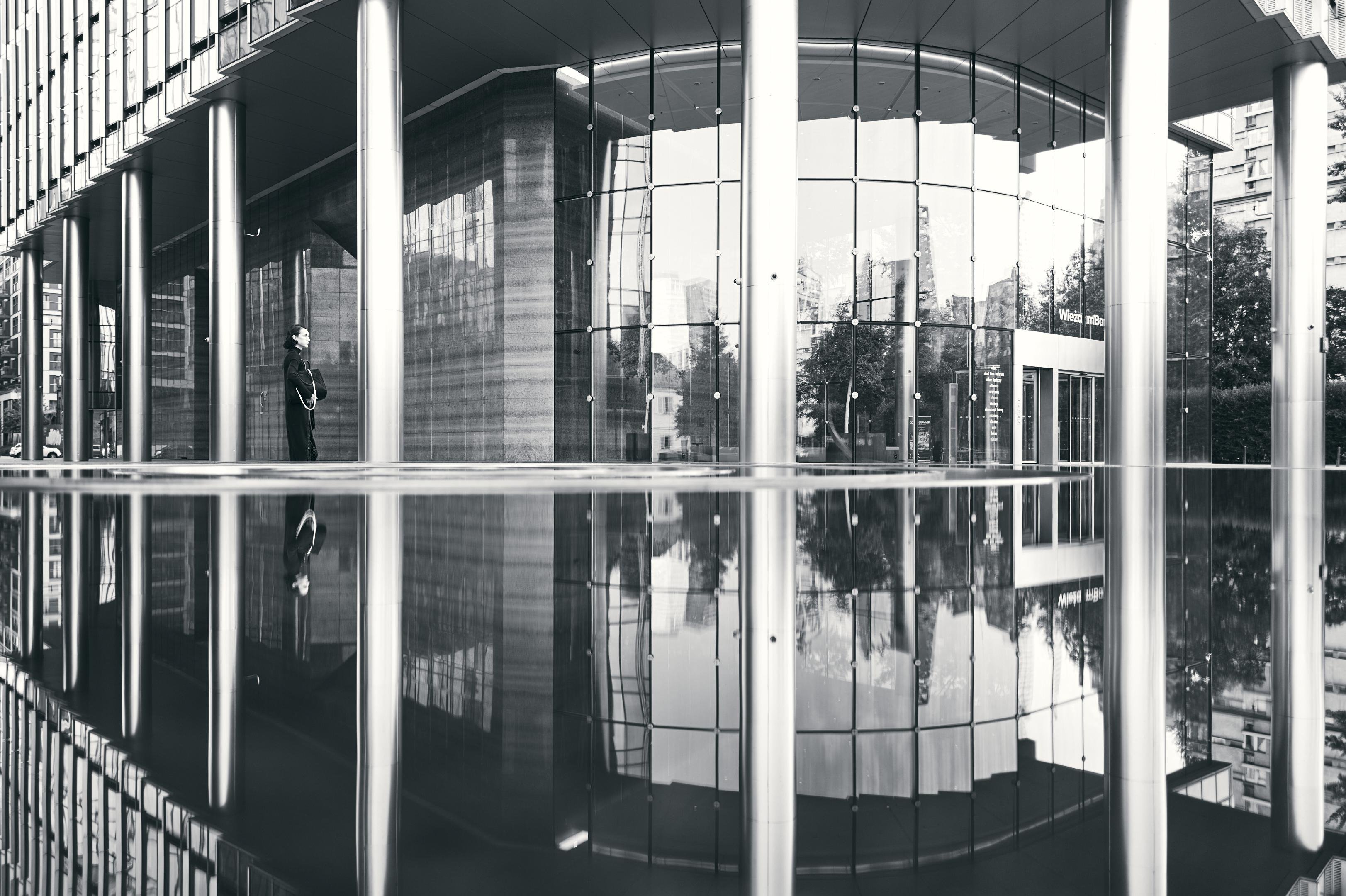 Black-and-white photo of a modern glass building entrance with tall columns, reflected in a still pool of water; a person stands near the left side under the overhang.