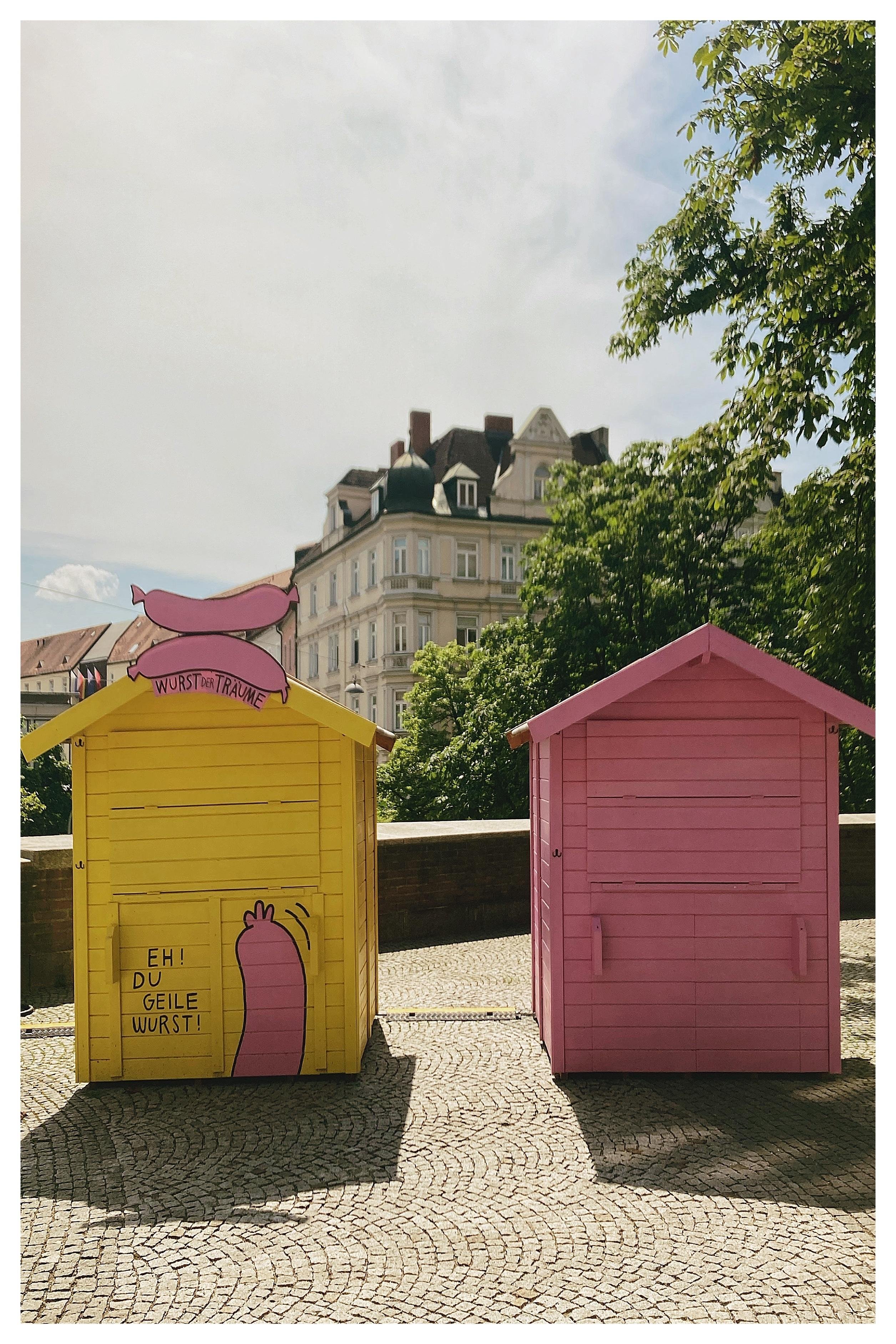 Two small wooden kiosks on a cobblestone plaza, one yellow with a pink sausage sign and German text and one solid pink, with trees and a multi-story historic building in the background under a bright sky.