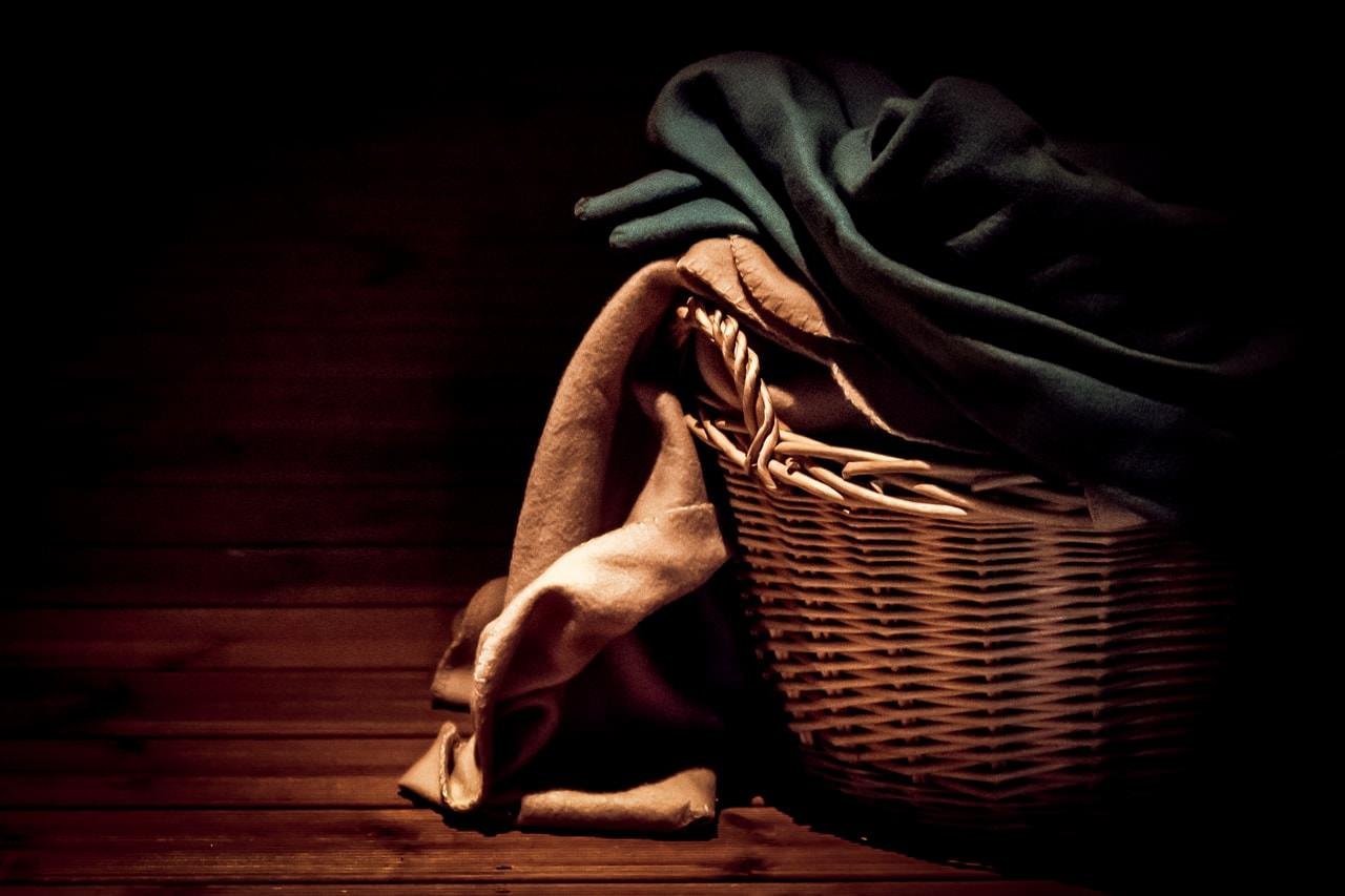 Wicker basket on a wooden floor, filled with folded green and beige blankets draping over the sides. Dim lighting emphasizes shadows, creating a cozy, rustic atmosphere.