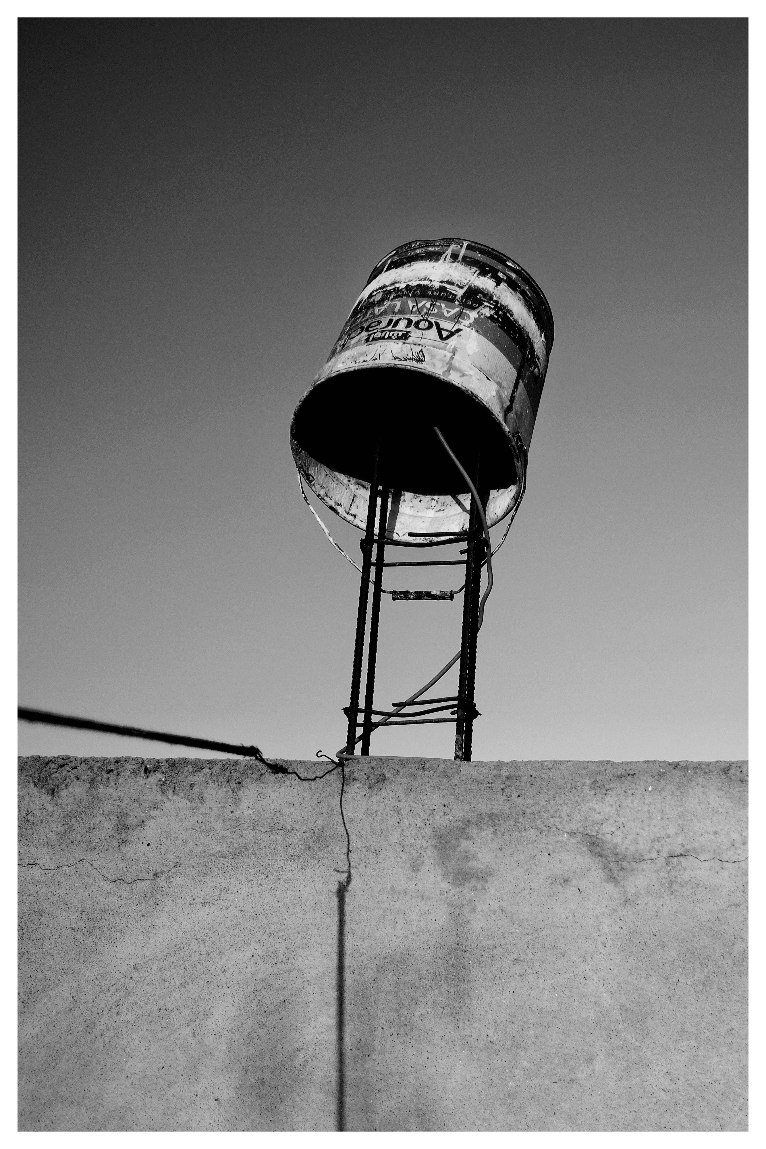 Black-and-white low-angle view of a weathered cylindrical metal tank on a narrow steel stand above a concrete wall, set against a clear sky with a cable running along the wall.