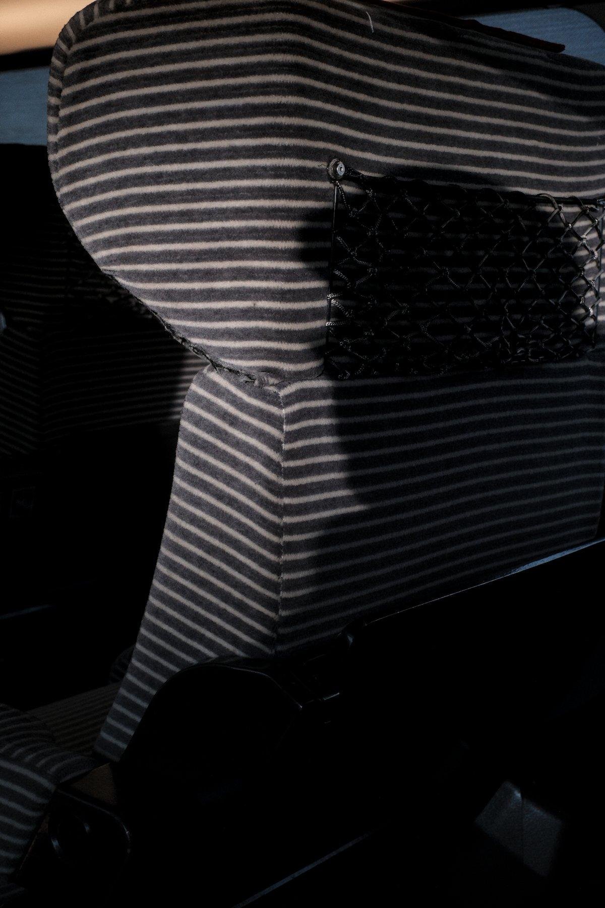 Photo verticale. Sur le dos d’un siège de train aux rayures horizontales, est projetée une ombre de la tête d’une personne portant un beret et des lunettes