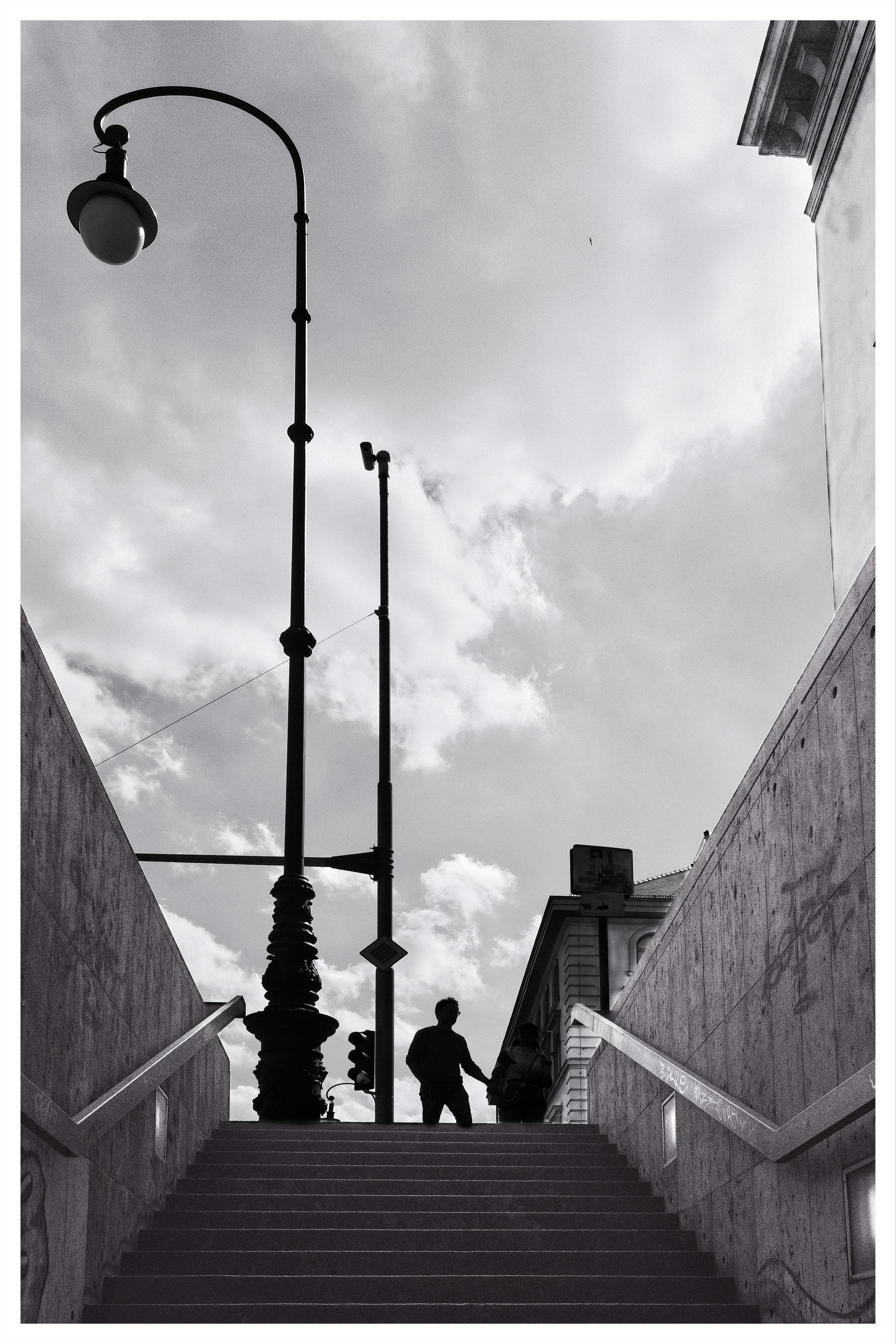 Black-and-white low-angle view from the bottom of a stairway leading up to street level, with concrete walls and handrails on both sides, a tall curved streetlamp and pole against a cloudy sky, and two silhouetted people at the top near a building edge.